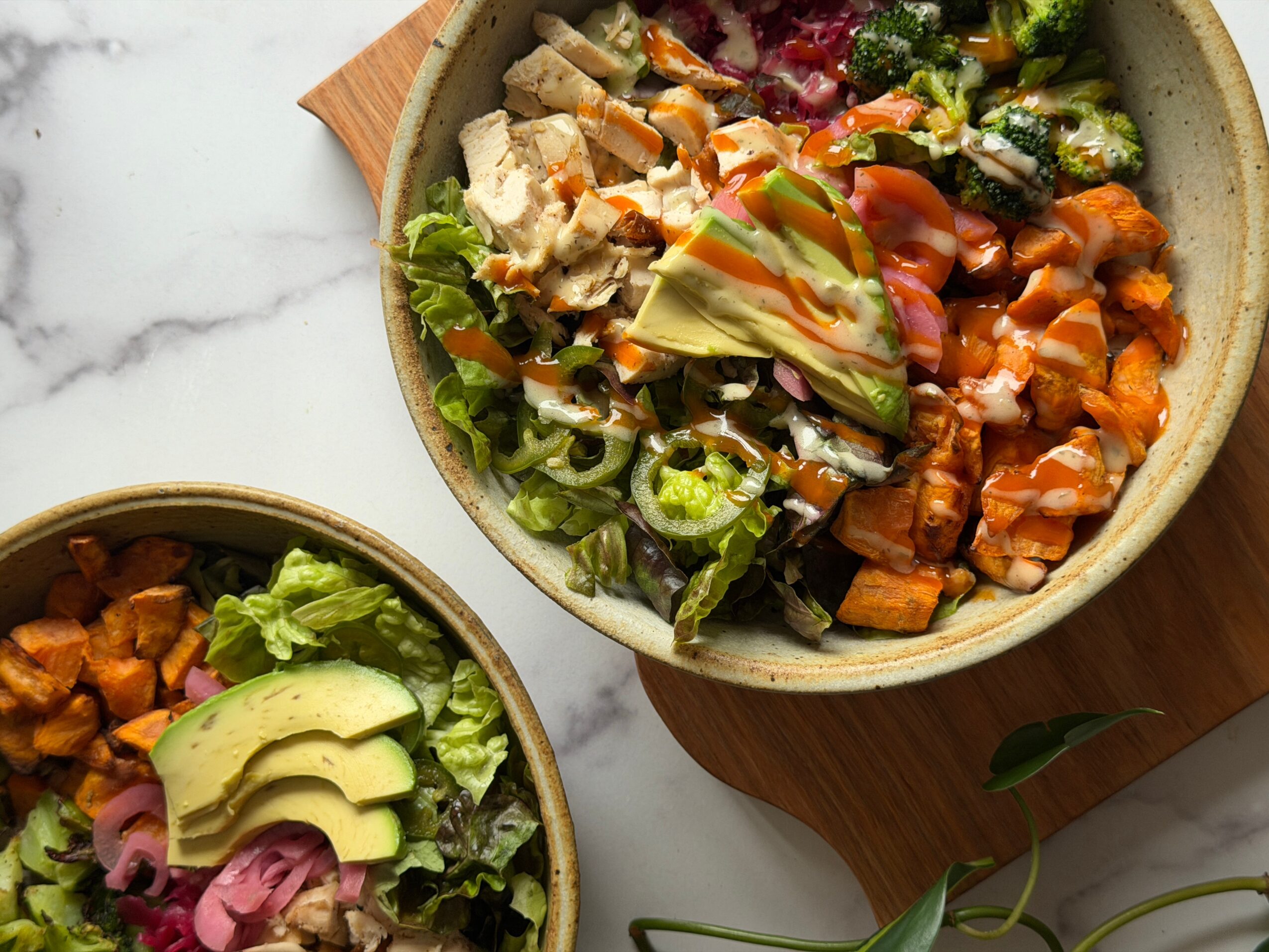 Two bowls of salad with vegetables, avocado, and chicken on a white surface, one on a wooden board.