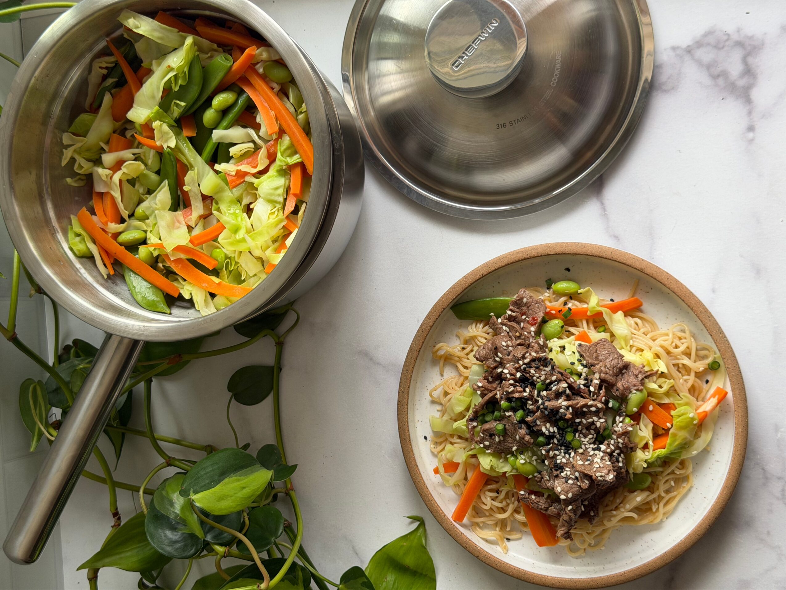 A bowl of cooked noodles with vegetables and meat, a pot with salad, and a lid on a marble surface.