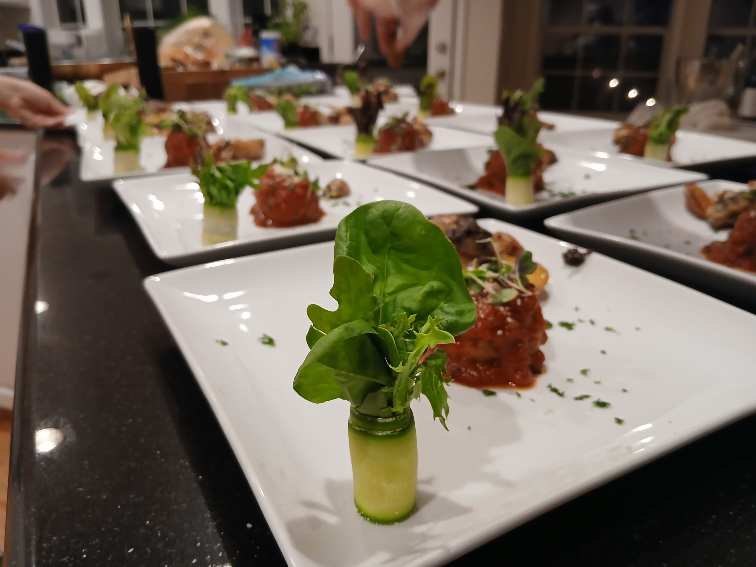 Multiple white plates with food, focus on a plate with a green lettuce leaf and a small rolled vegetable, in a dining setting.