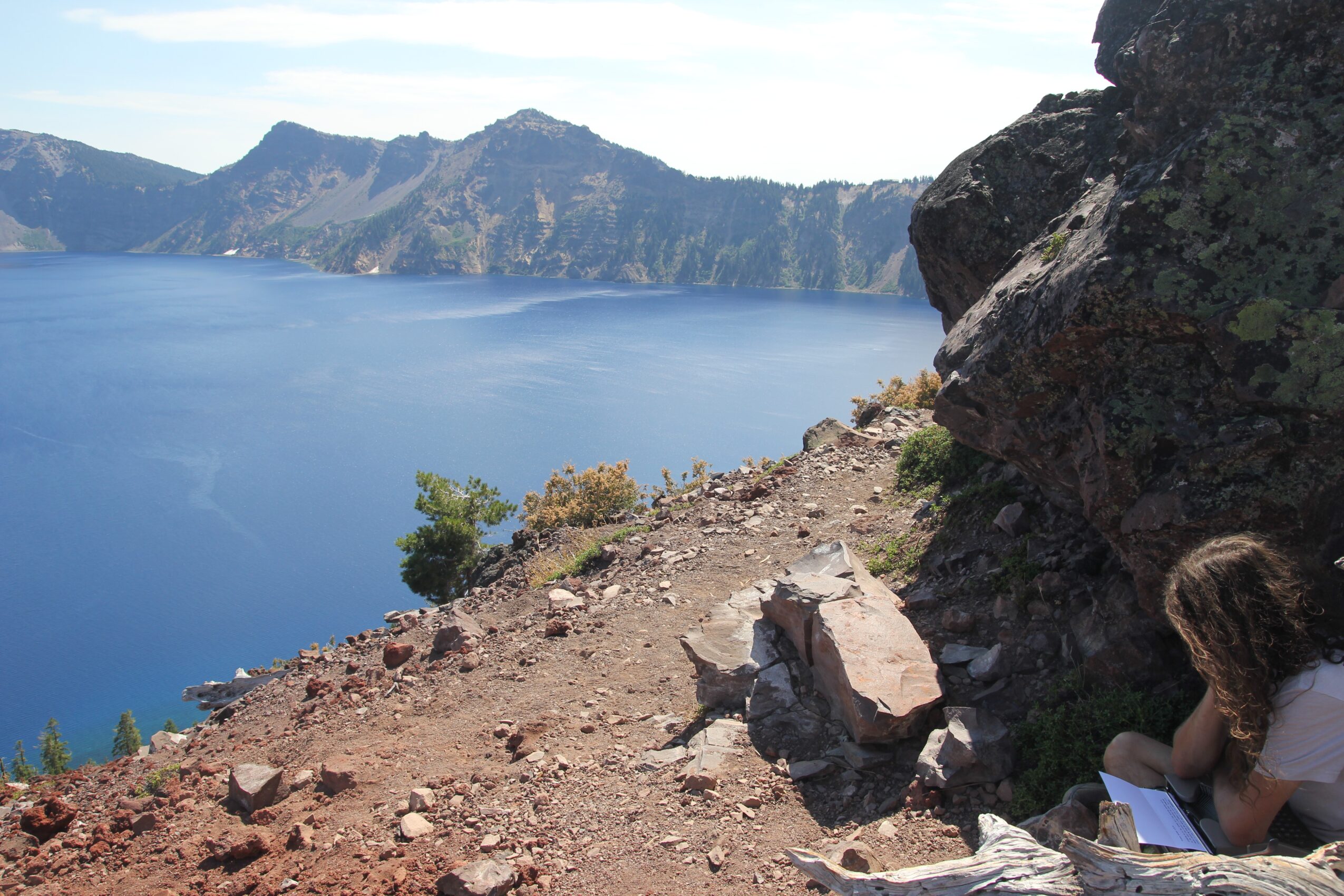 Person sitting on rocky ground near a large rock, overlooking a lake with mountains in the background, writing or drawing in a notebook.