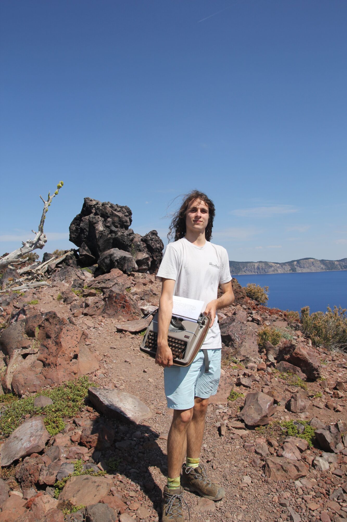 Person standing on rocky terrain near the coast, holding a tablet, with a clear blue sky and ocean in the background.