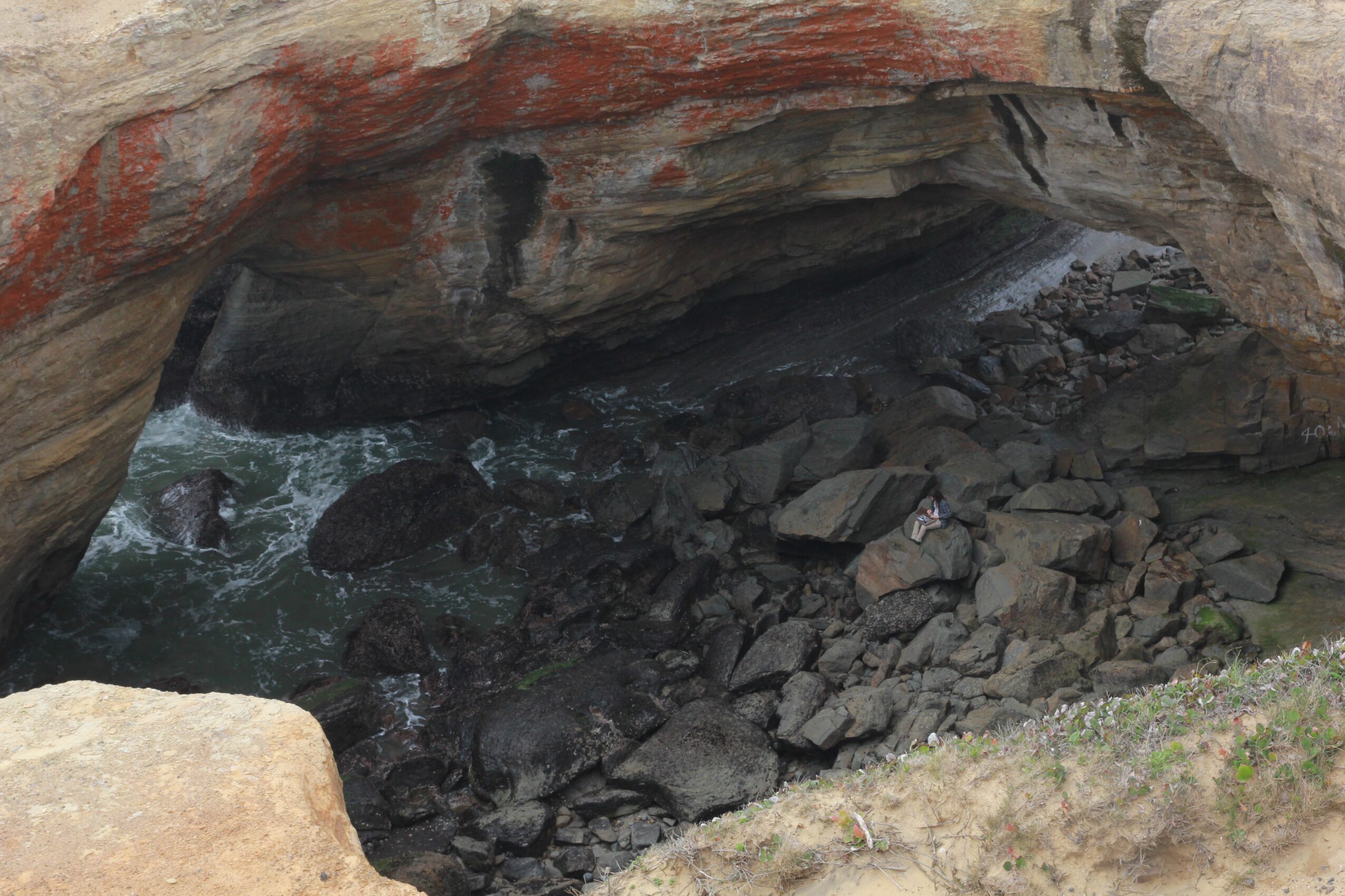 View of a rocky cave opening with water and rocks inside, seen from outside the cave entrance.