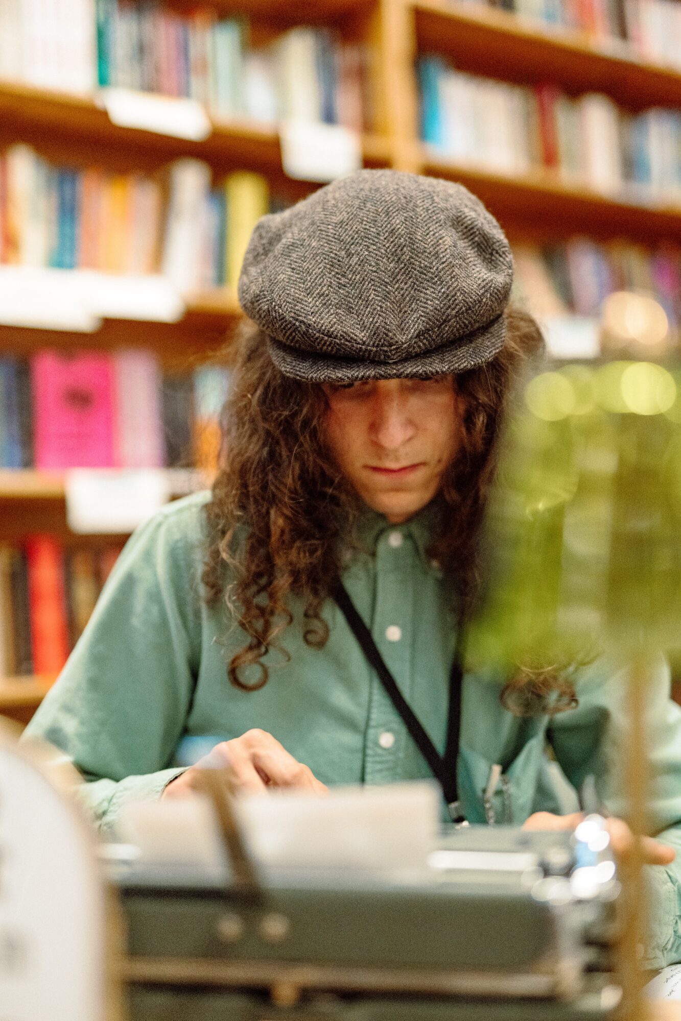 Person with long curly hair wearing a gray cap and green shirt, reading in a library with bookshelves in background.