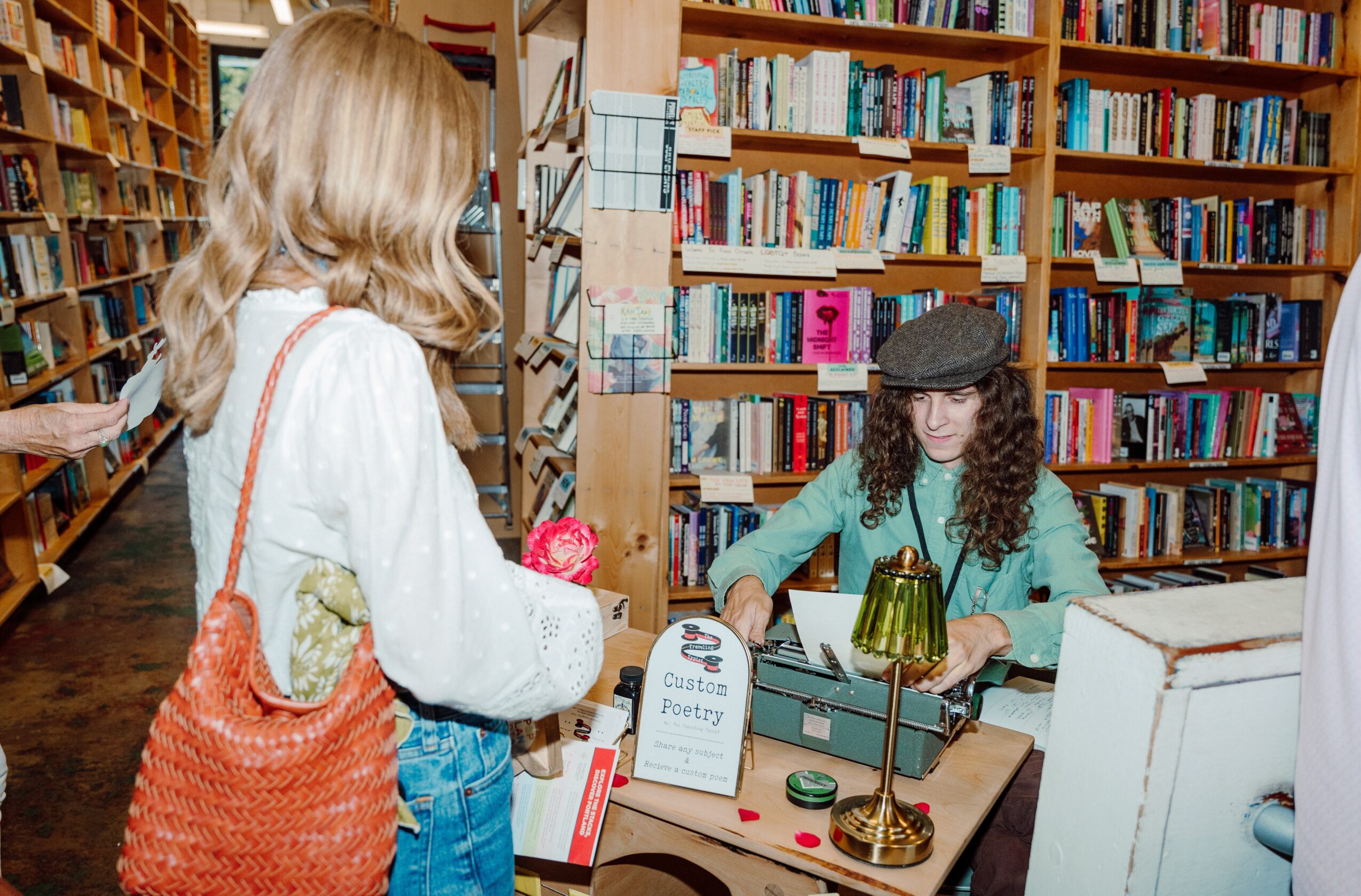 Two women in a bookstore, one browsing and the other sitting at a desk with books and a green lamp.