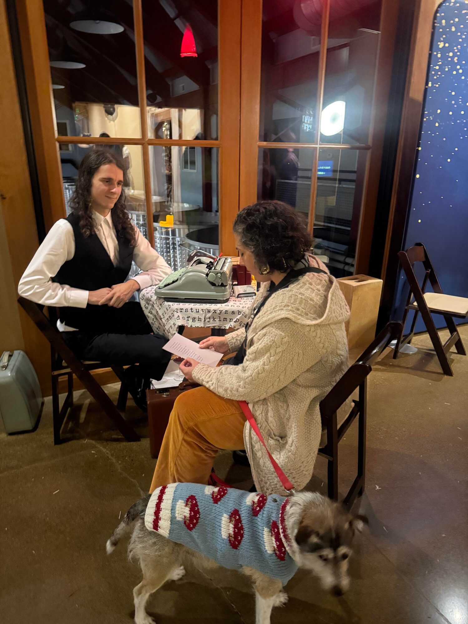Two women sit at a table in a cozy indoor setting, one reading a book, with a dog wearing a sweater nearby.
