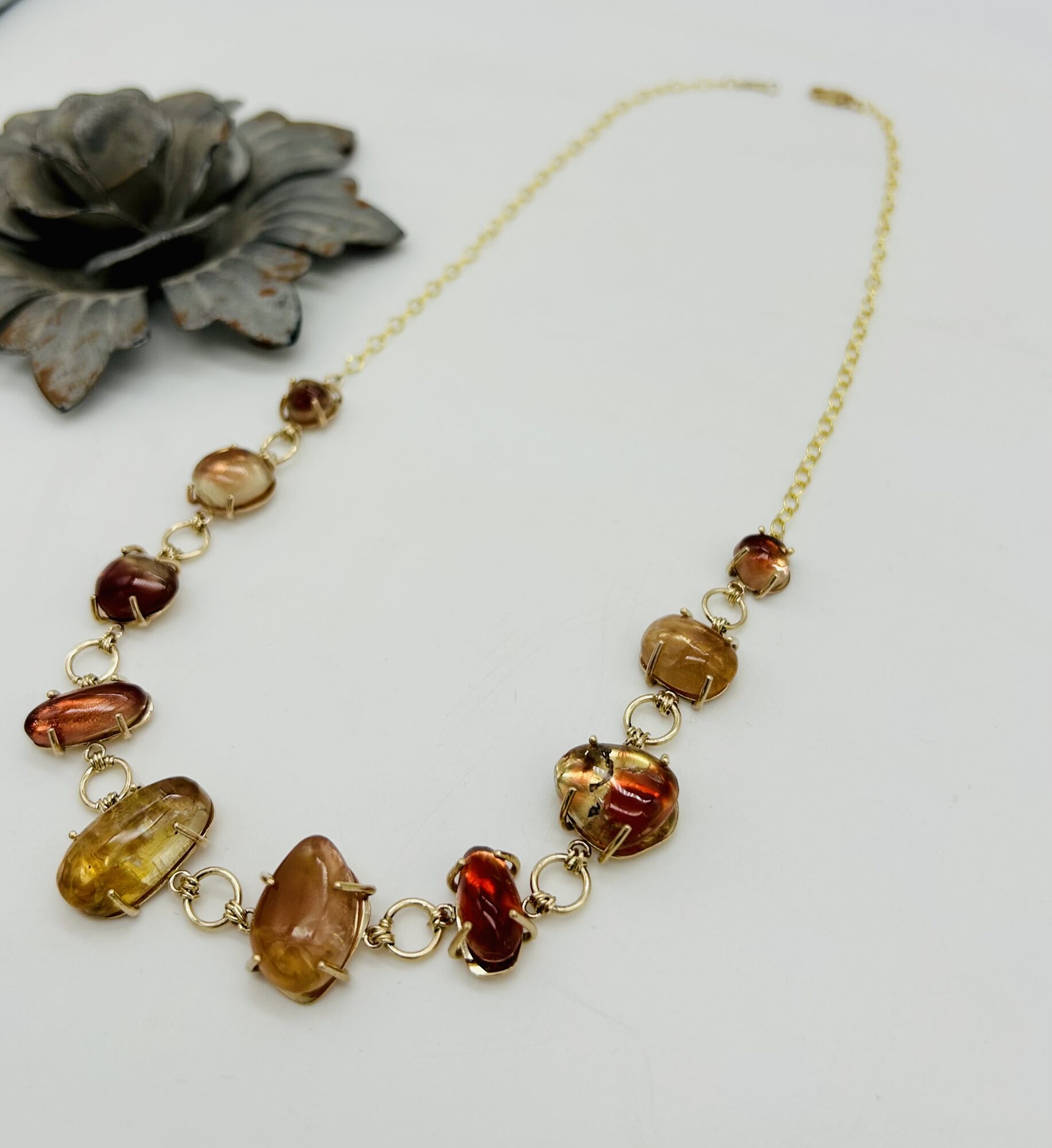 Gold chain necklace with large amber and red beads, placed on a white surface with a dark flower decoration nearby.