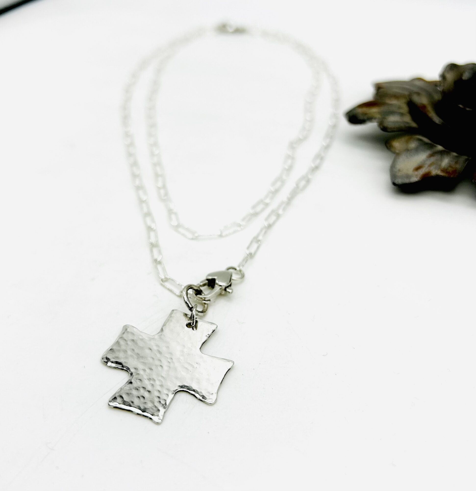Silver cross pendant necklace with chain, placed on a white surface with a dark flower to the right.
