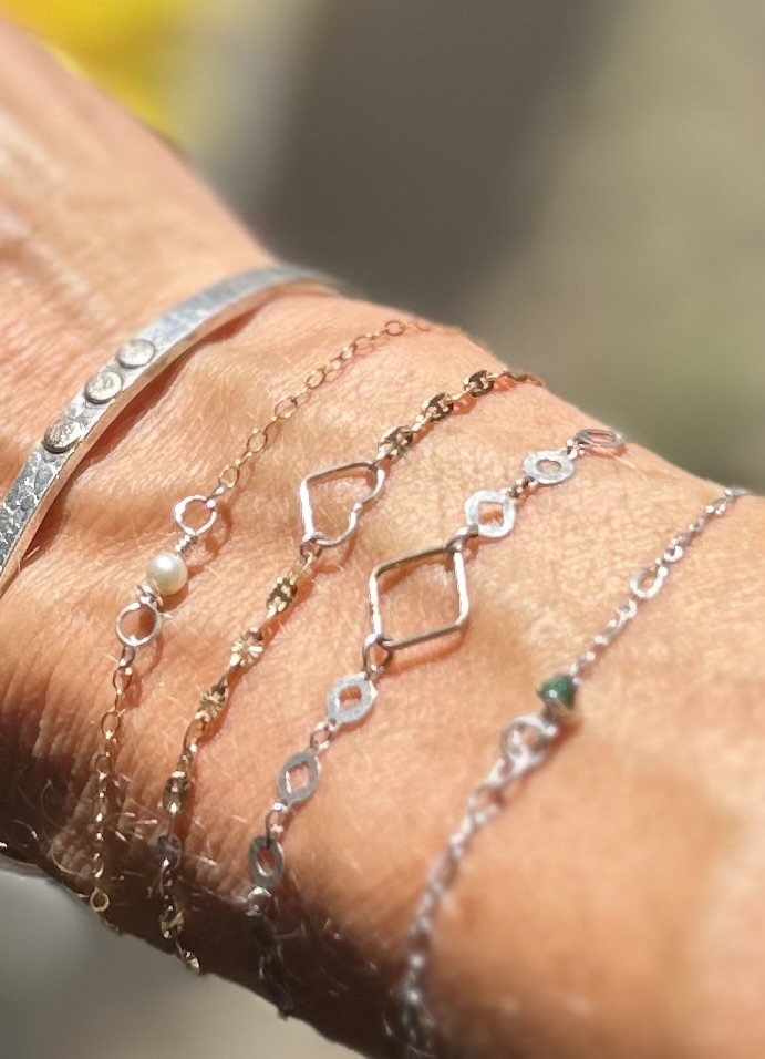 Multiple delicate bracelets with various chain designs and small charms on a person's wrist, sunlight highlighting the jewelry.