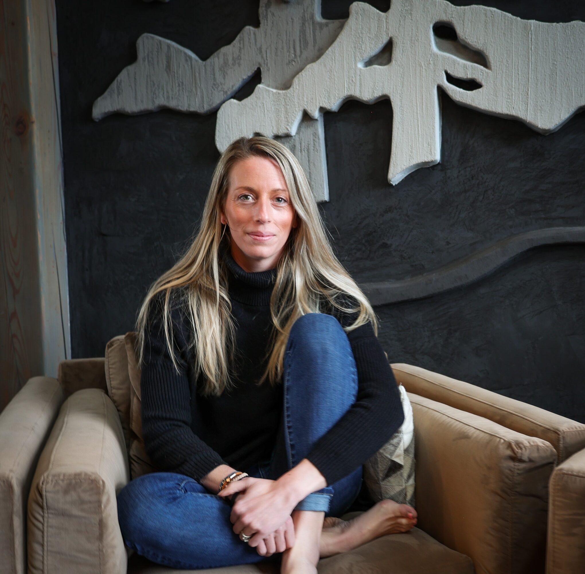 Woman with long blonde hair sitting on a beige sofa, wearing a black turtleneck and blue jeans, in a room with dark wall art.