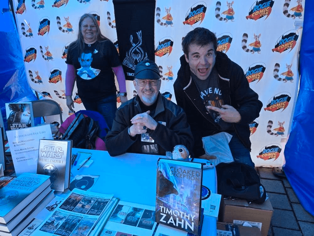 Three people at a table with books and promotional materials, smiling and posing for the camera, with a patterned backdrop behind them.