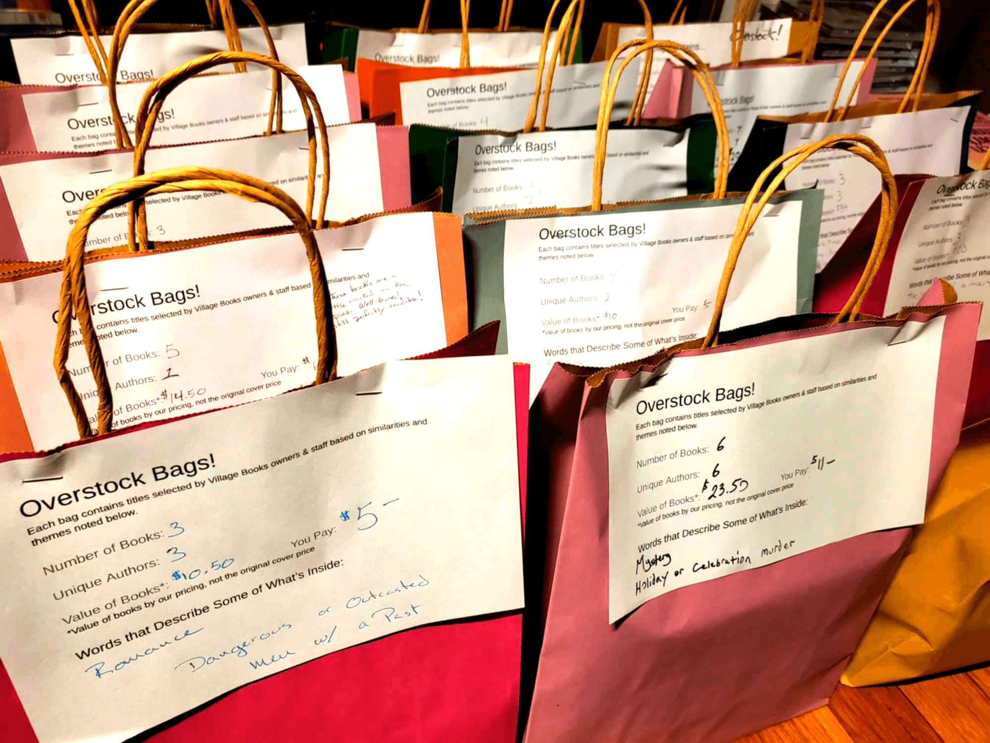 Multiple colorful paper shopping bags with handles, some with handwritten notes, arranged closely together.