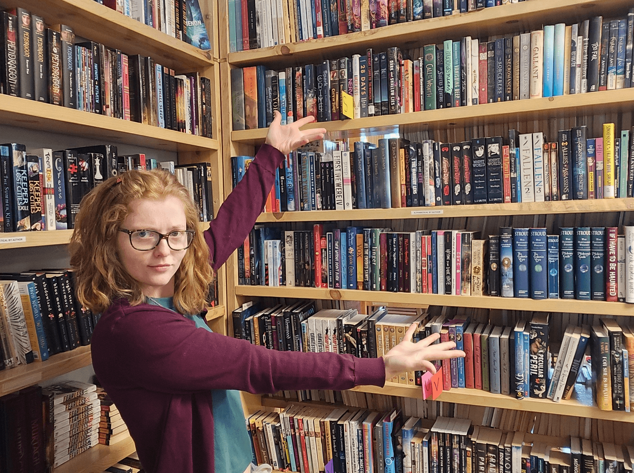 Woman with glasses and curly hair reaching for books on a bookshelf in a library.