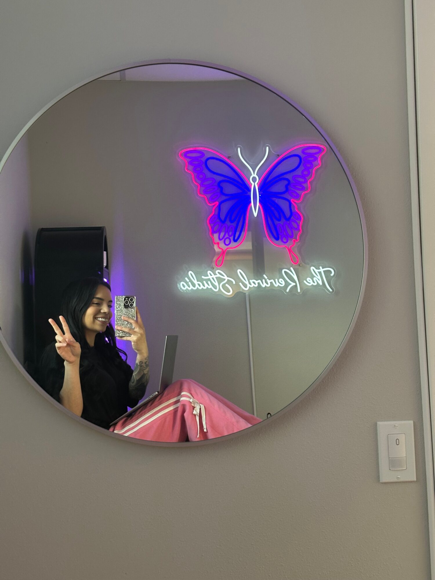 Round mirror with neon butterfly and text, person taking photo reflected in mirror, wall with electrical outlet, pink object partially visible