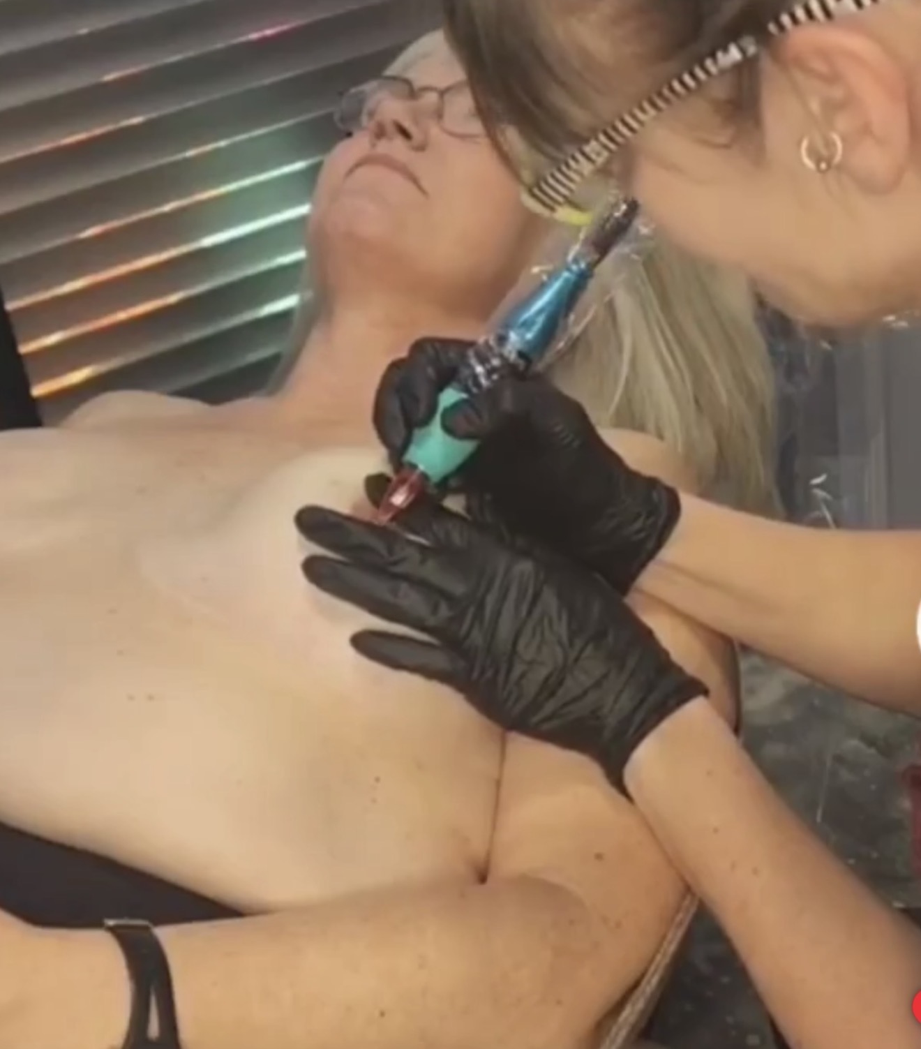 Person receiving a tattoo on their chest while lying down, with a tattoo artist using a tattoo machine. The person has glasses and blonde hair.