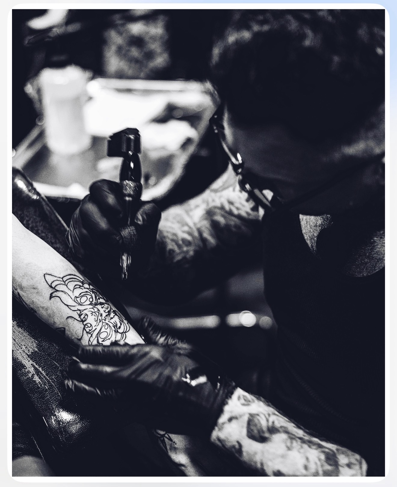 Tattoo artist working on a person's arm with tattoo equipment, wearing gloves and glasses, in a tattoo studio.
