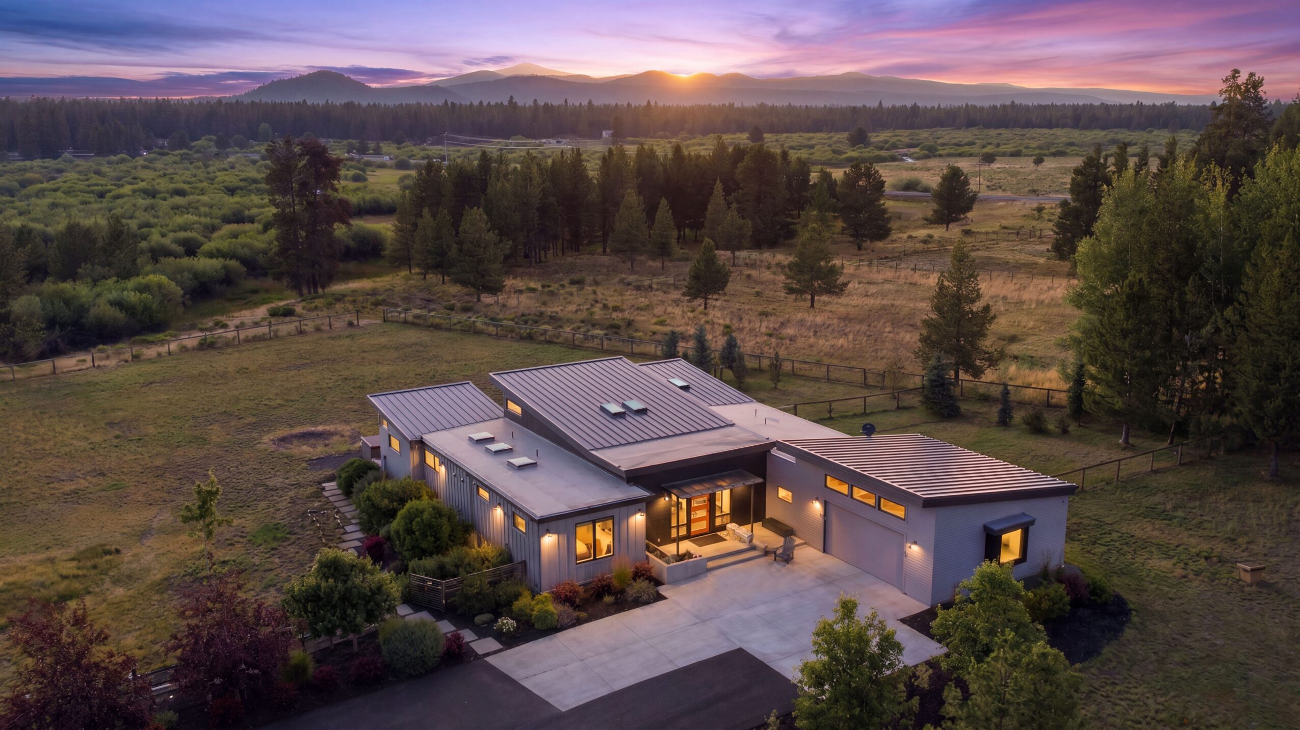 A modern house with multiple sections, surrounded by greenery and trees, under a sunset sky.