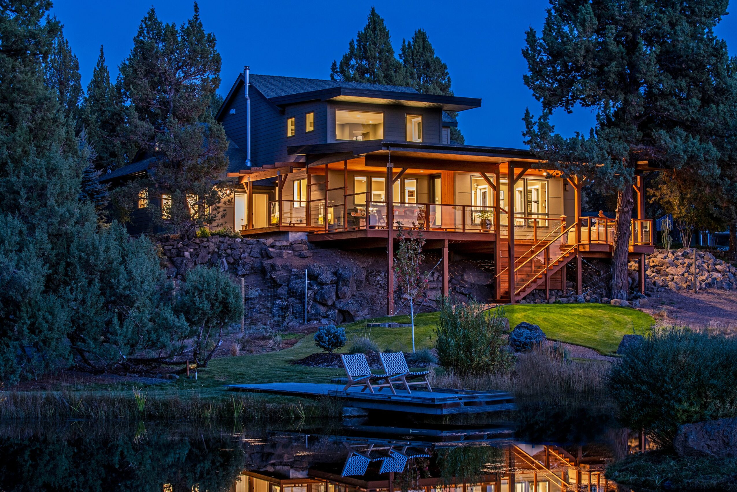 A house on a hill illuminated at dusk, with a deck overlooking a pond and trees, reflecting in the water.