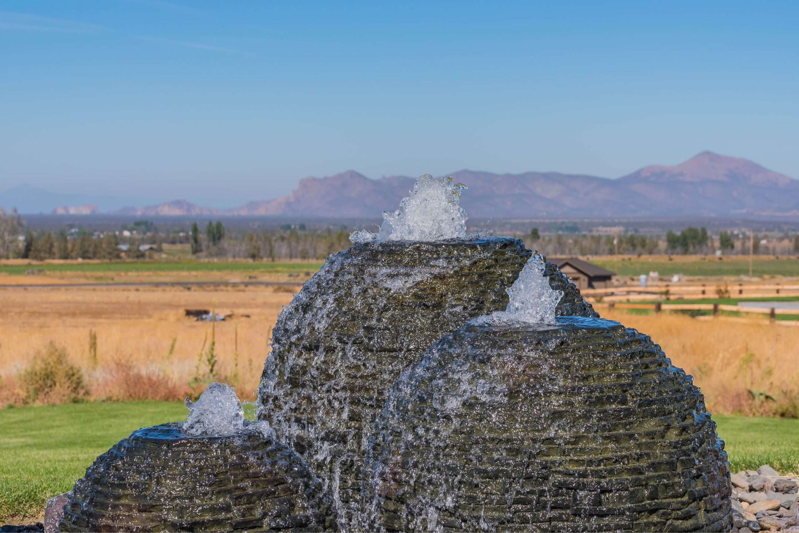 Fountain with water splashing, set against a landscape with fields and mountains in the background.