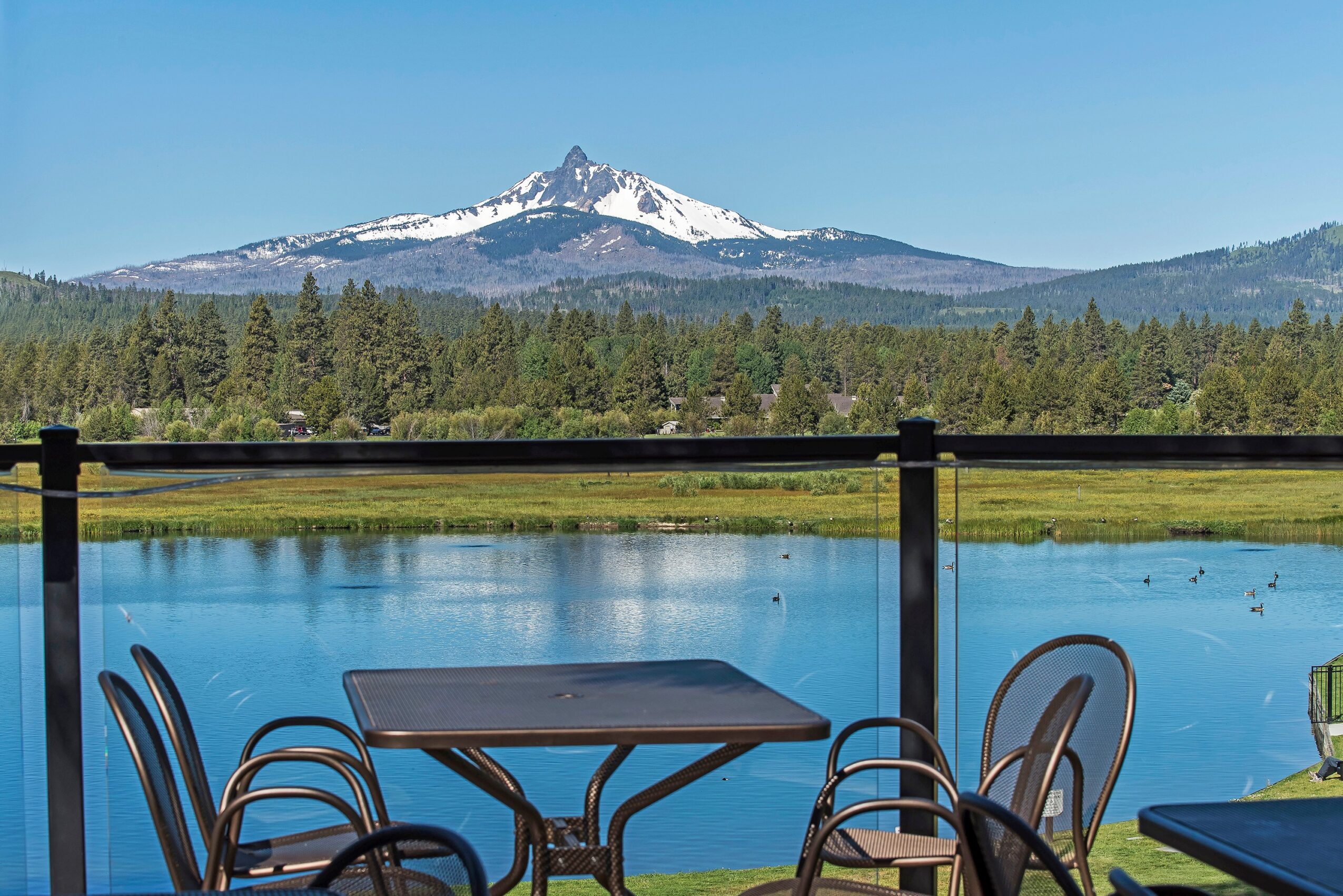 Outdoor scene with a table and chairs by a lake, mountain with snow in background, clear blue sky.