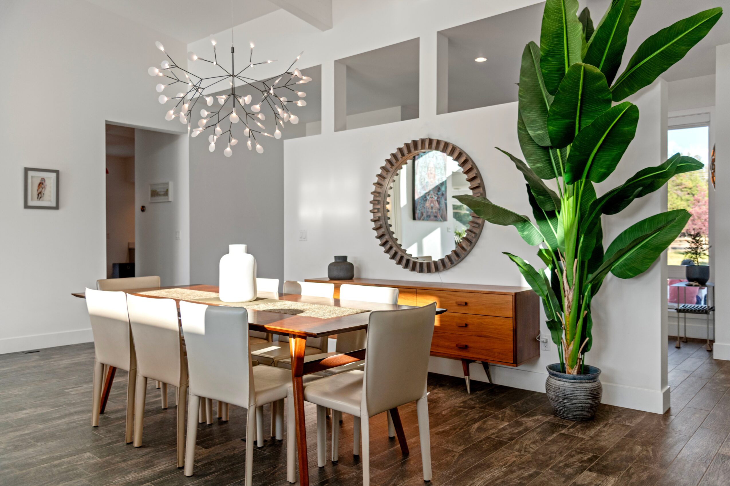 Dining room with a wooden table, white chairs, a large green plant, a round mirror, and a modern chandelier.