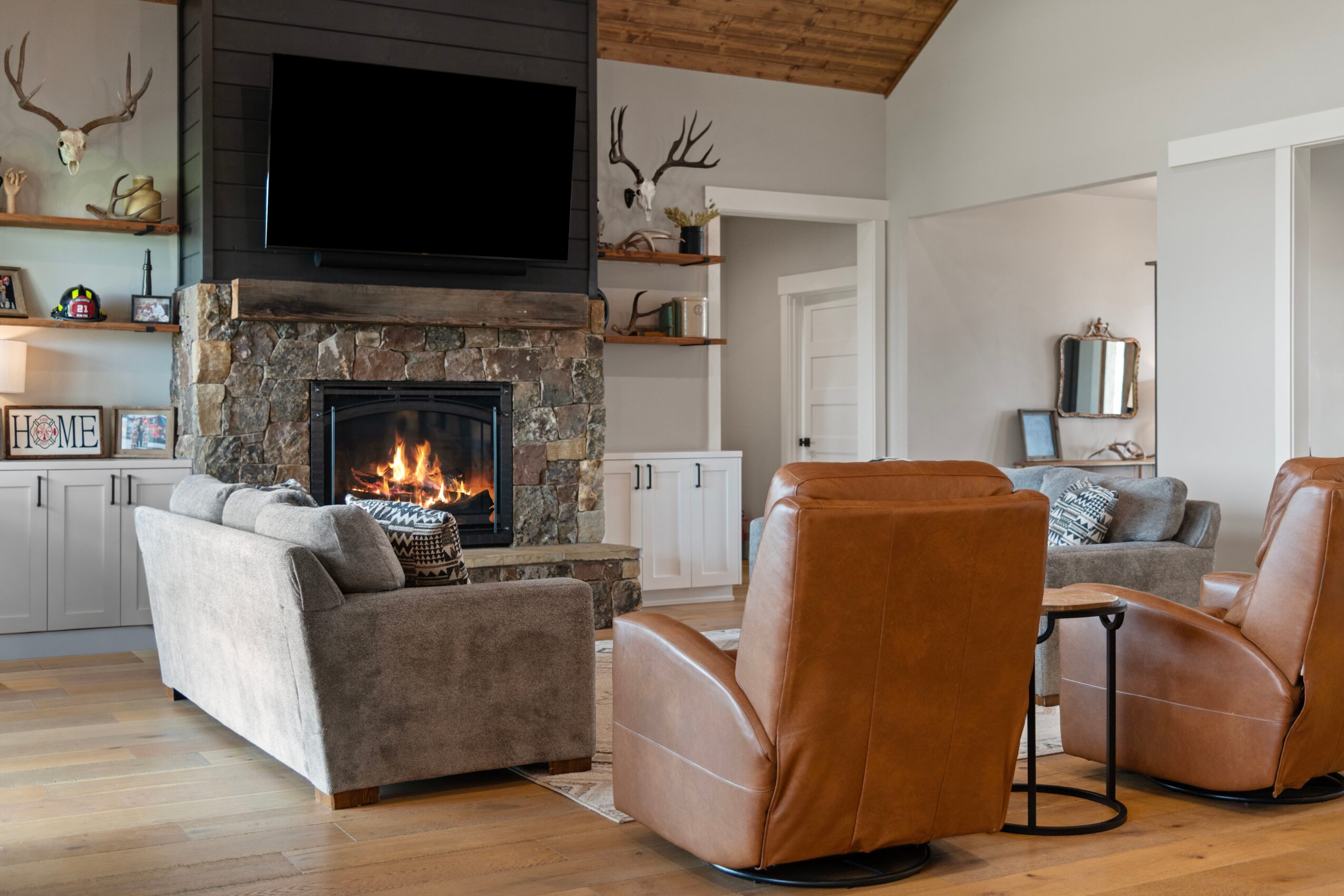 Living room with a stone fireplace, TV above, gray sofa, two brown leather chairs, and wooden ceiling beams.