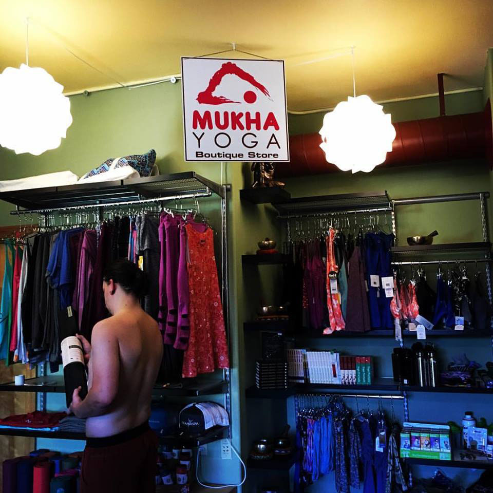 Clothing store interior with racks of colorful clothes, a person browsing, and a sign reading 'MUKHA YOGA Boutique Store'.