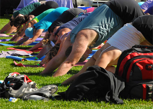 Group of people practicing yoga outdoors on grass, bending forward on mats, with bags and equipment nearby.