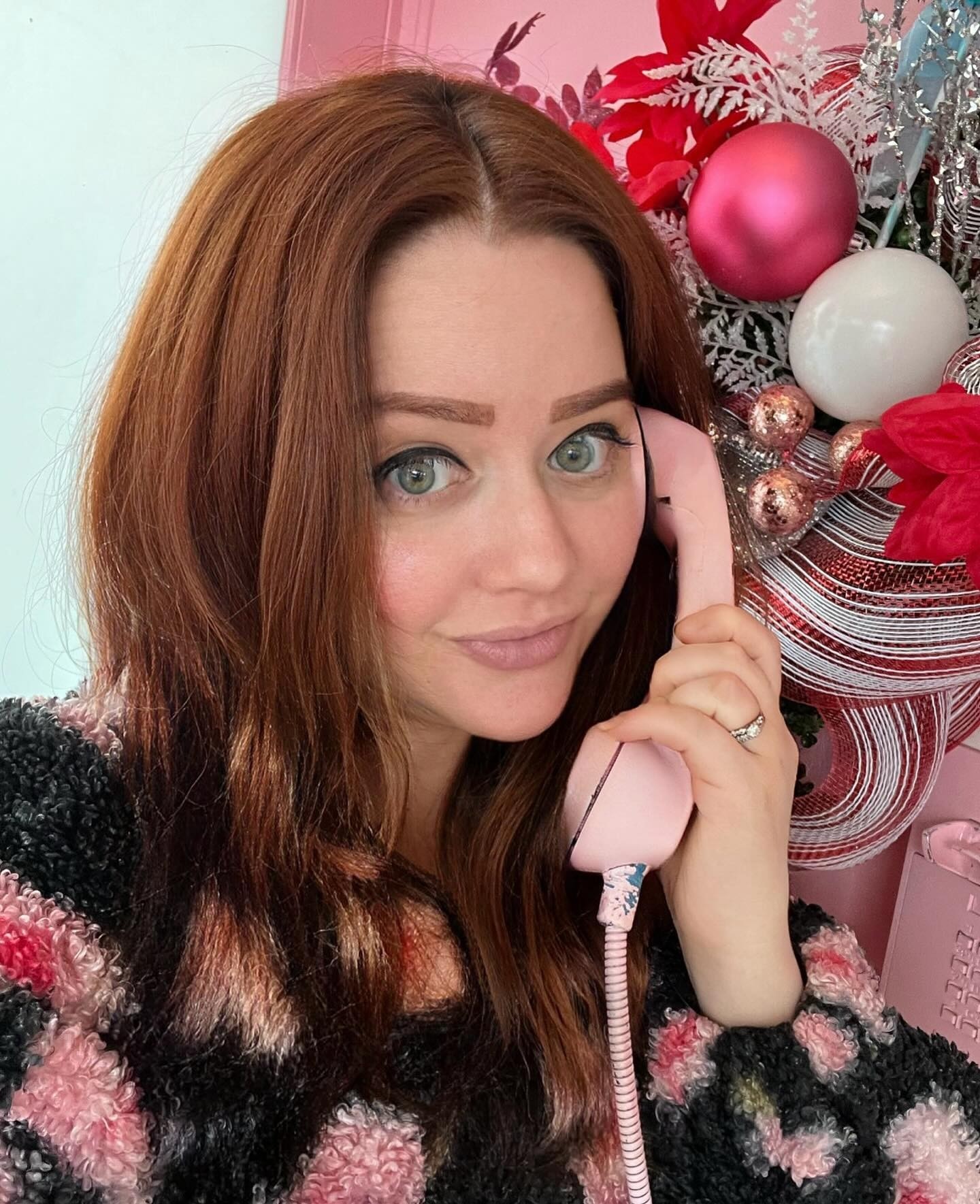 Woman with long red hair holding a pink phone, Christmas decorations in background, smiling softly.