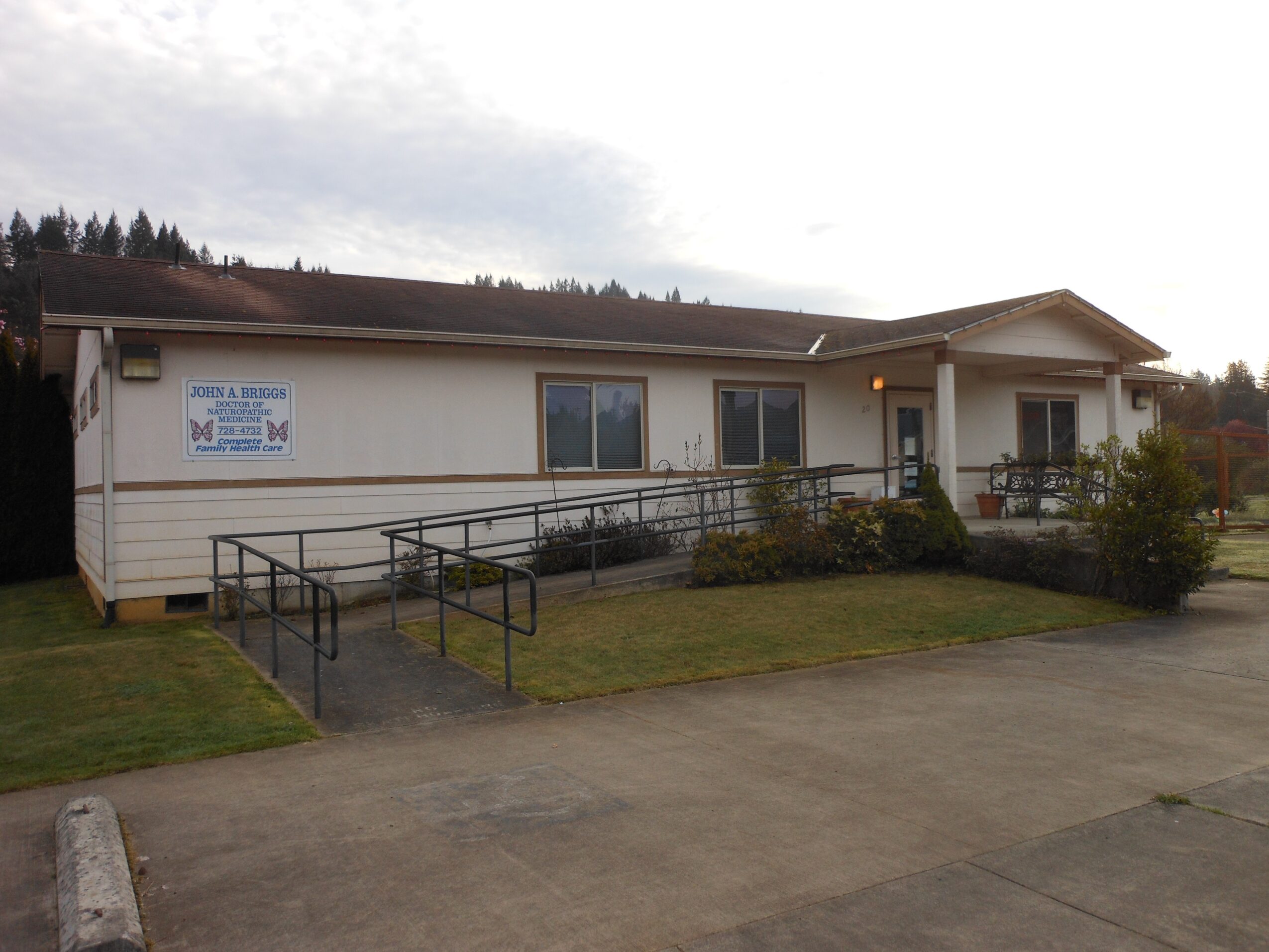 Single-story building with a ramp, windows, and a small porch, surrounded by grass and bushes, under a cloudy sky.