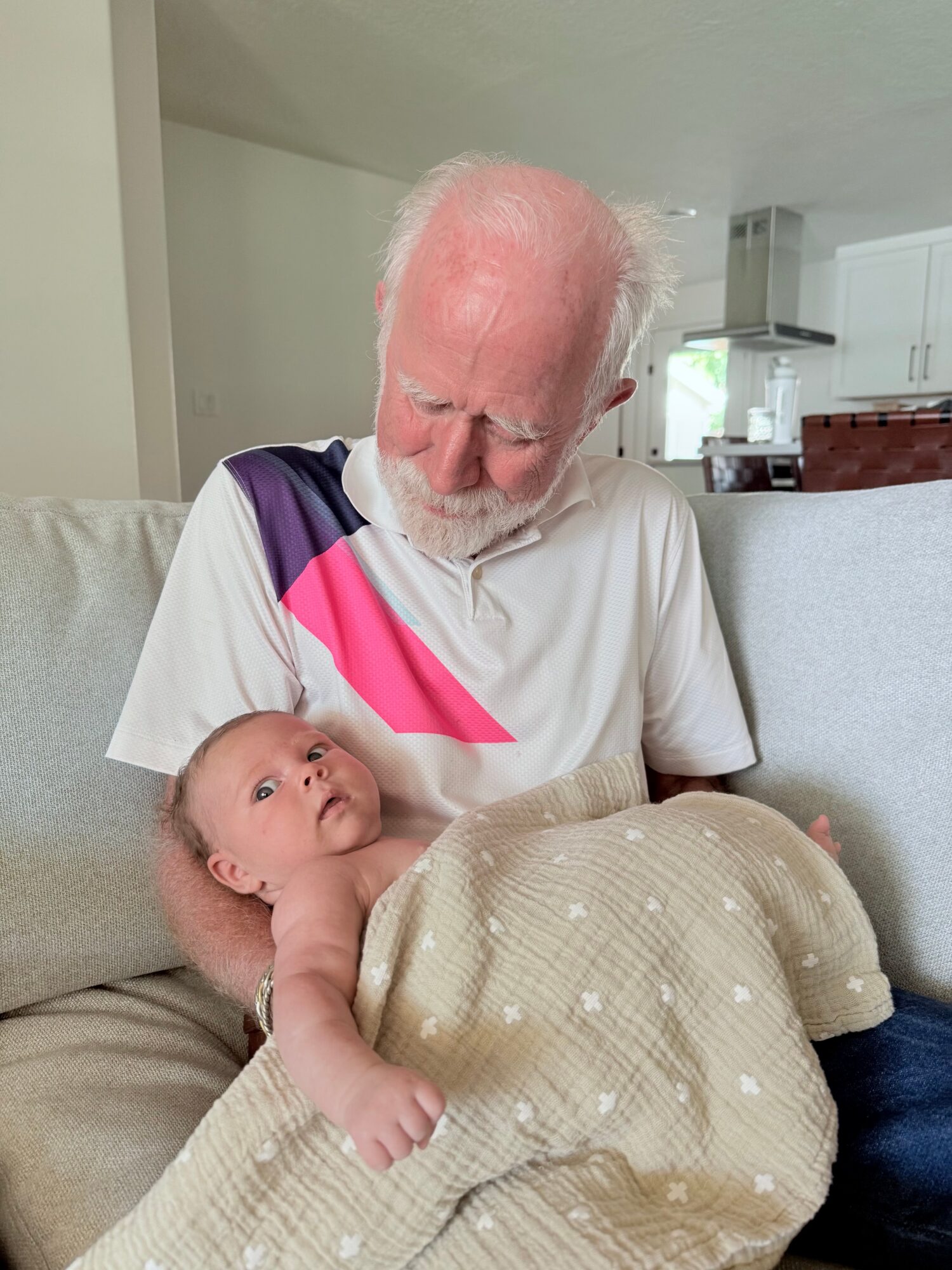 Elderly man with white hair and beard looking at a baby lying on a couch, wrapped in a beige blanket, in a bright room.