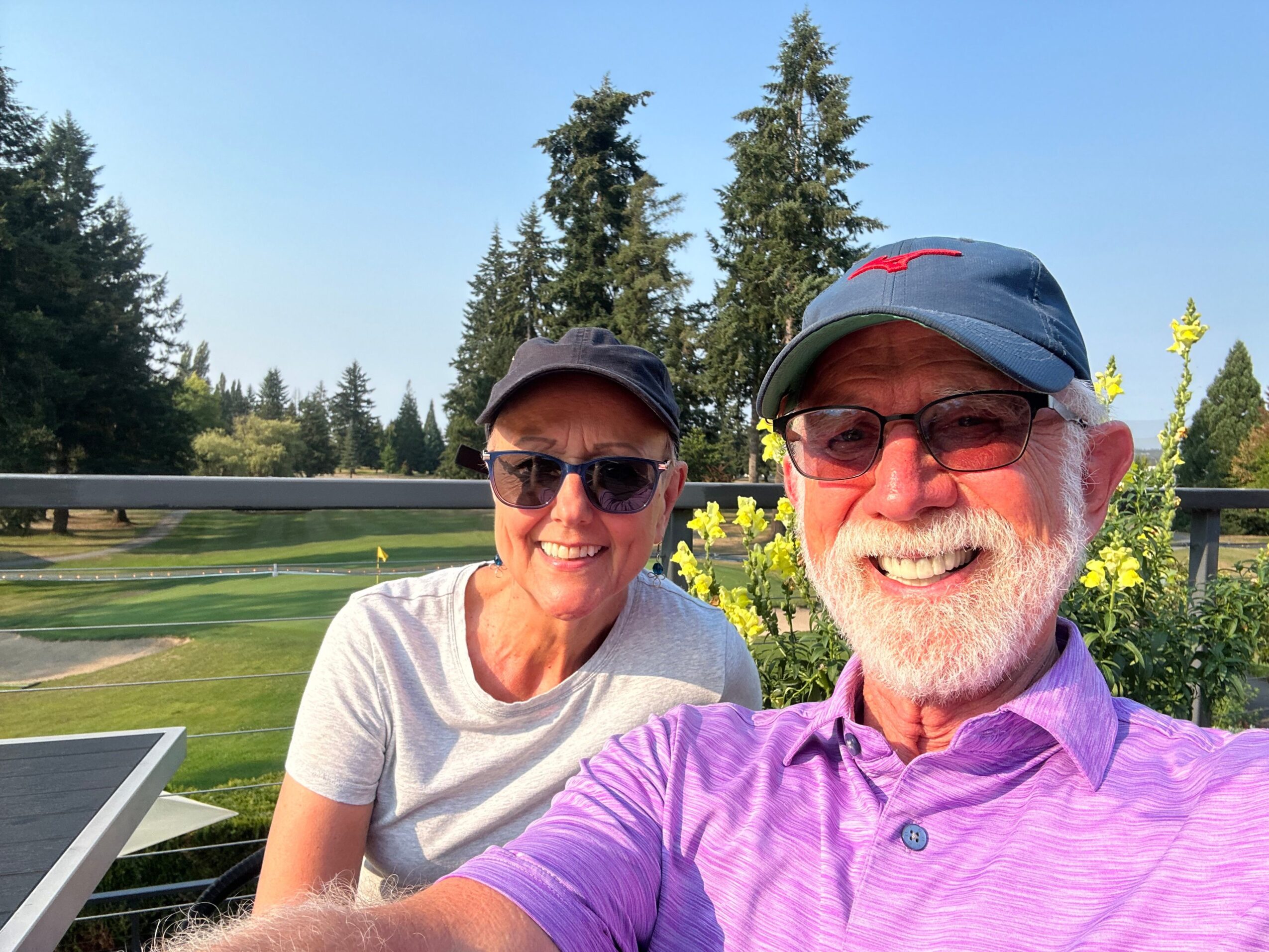 Two people smiling outdoors, one wearing a purple shirt and sunglasses, the other in a white shirt and sunglasses, with trees and sky in background.