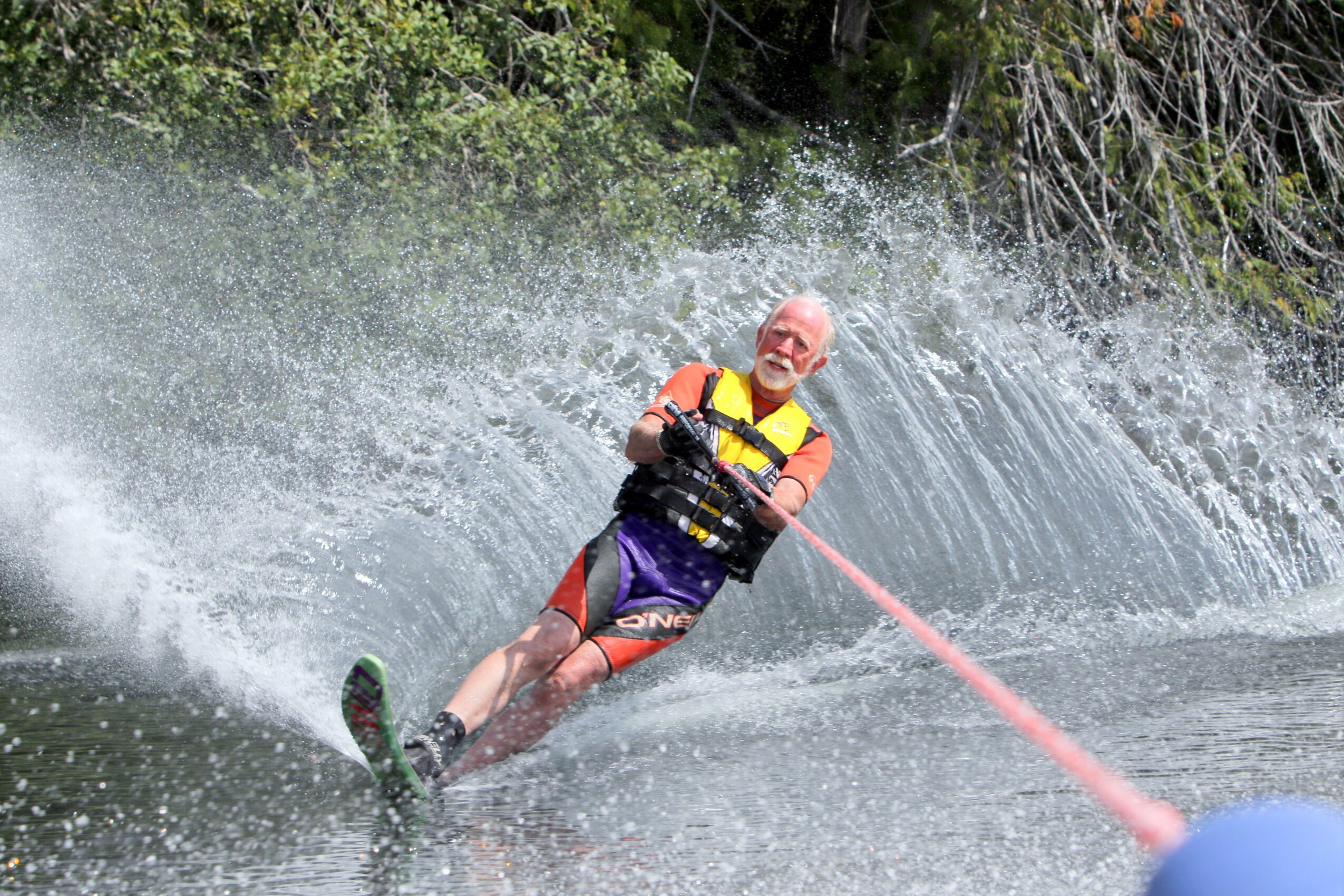 Person waterskiing on water, holding a pink tow rope, wearing a life jacket and sunglasses, with trees in background.