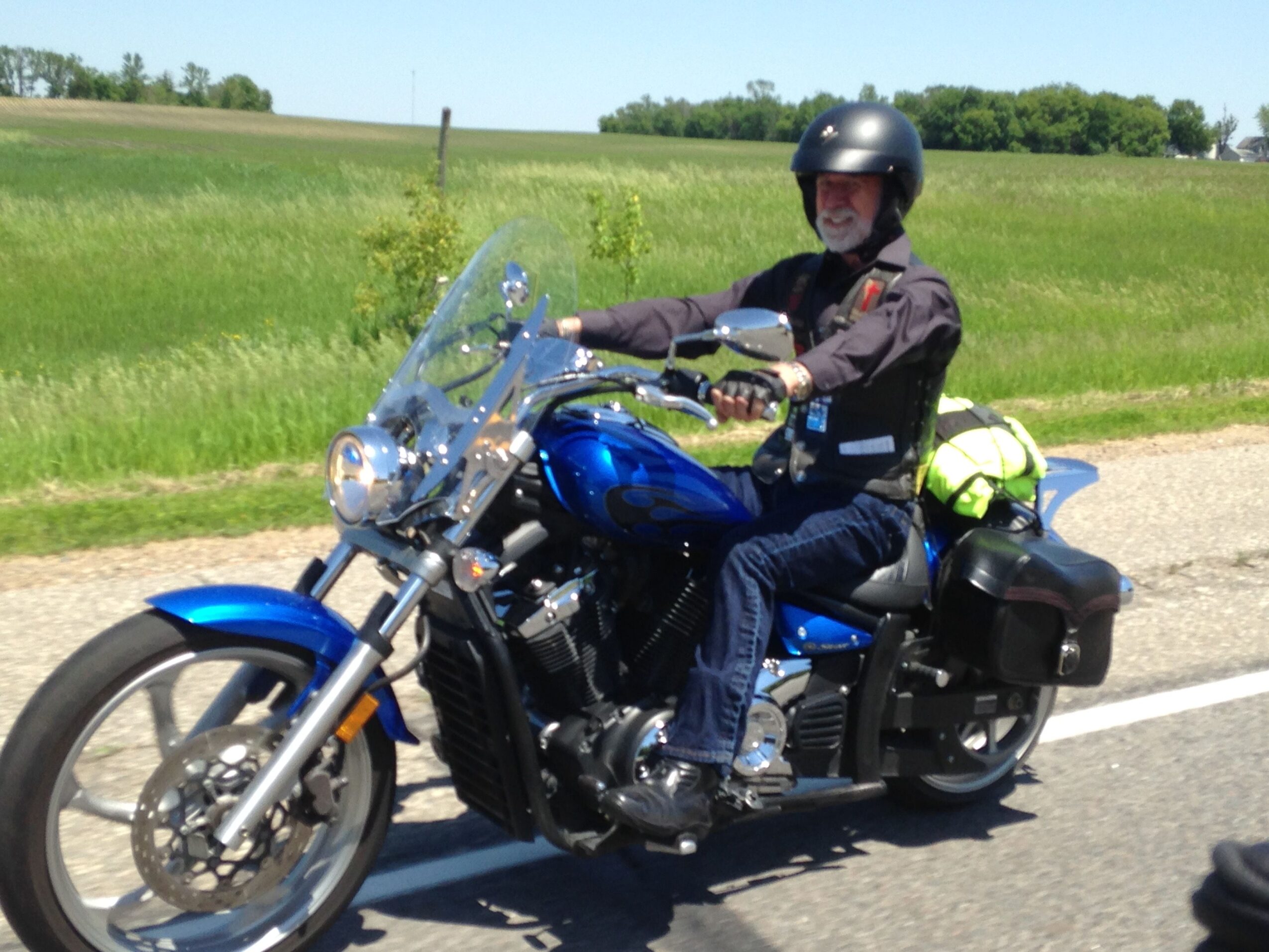 Motorcycle parked on grass with rider in black helmet and gear, blue bike, and clear windshield, landscape background.