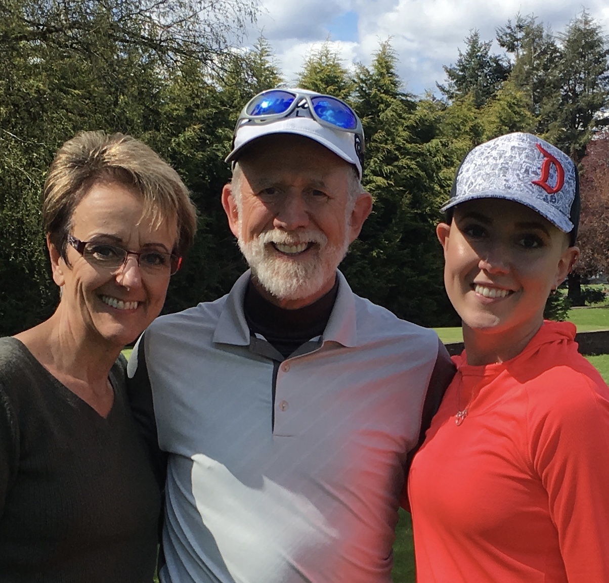 Three people smiling outdoors with trees and sky in background.