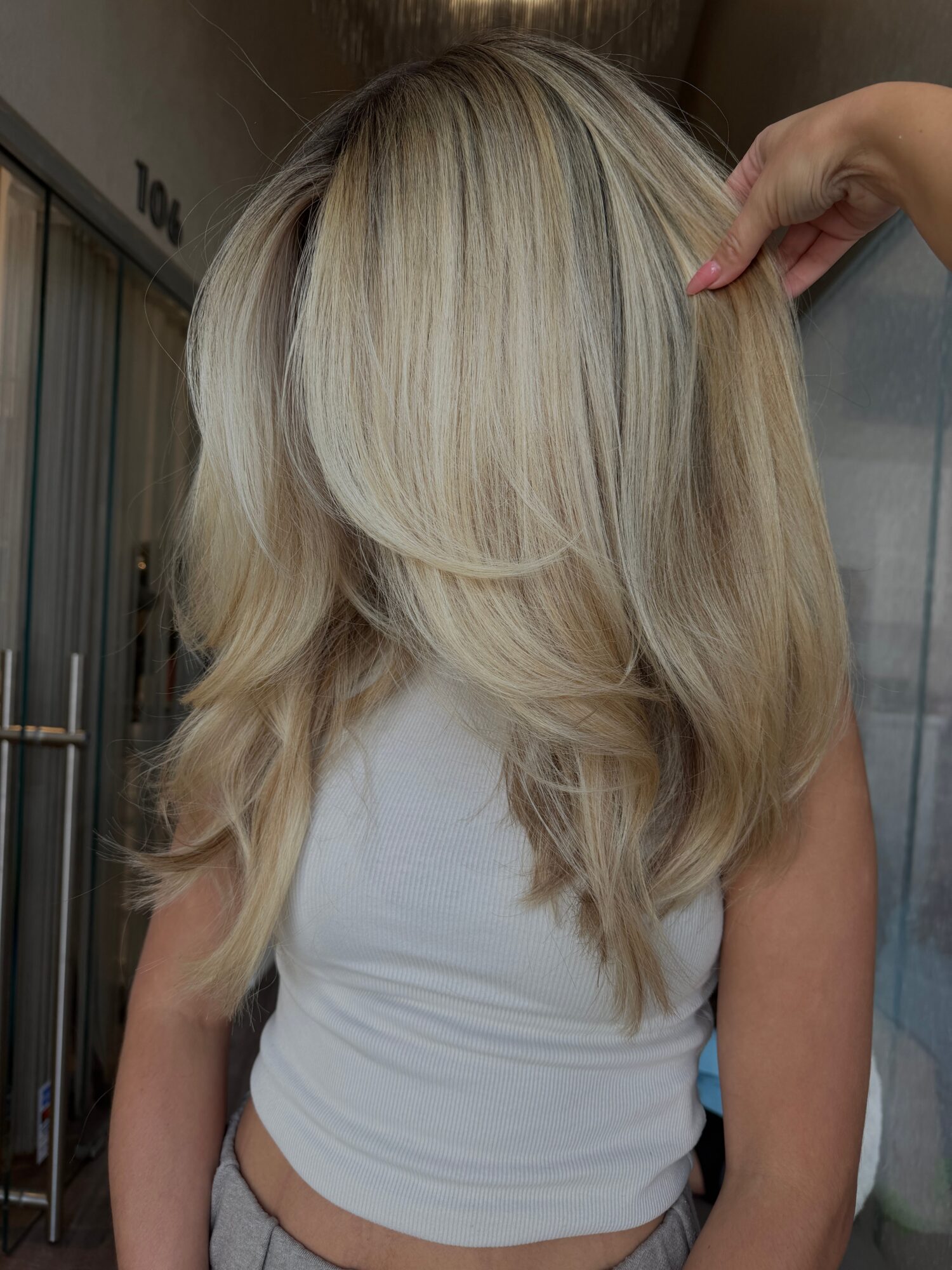 Person with long, layered blonde hair, wearing a white top, being held by another person's hand.