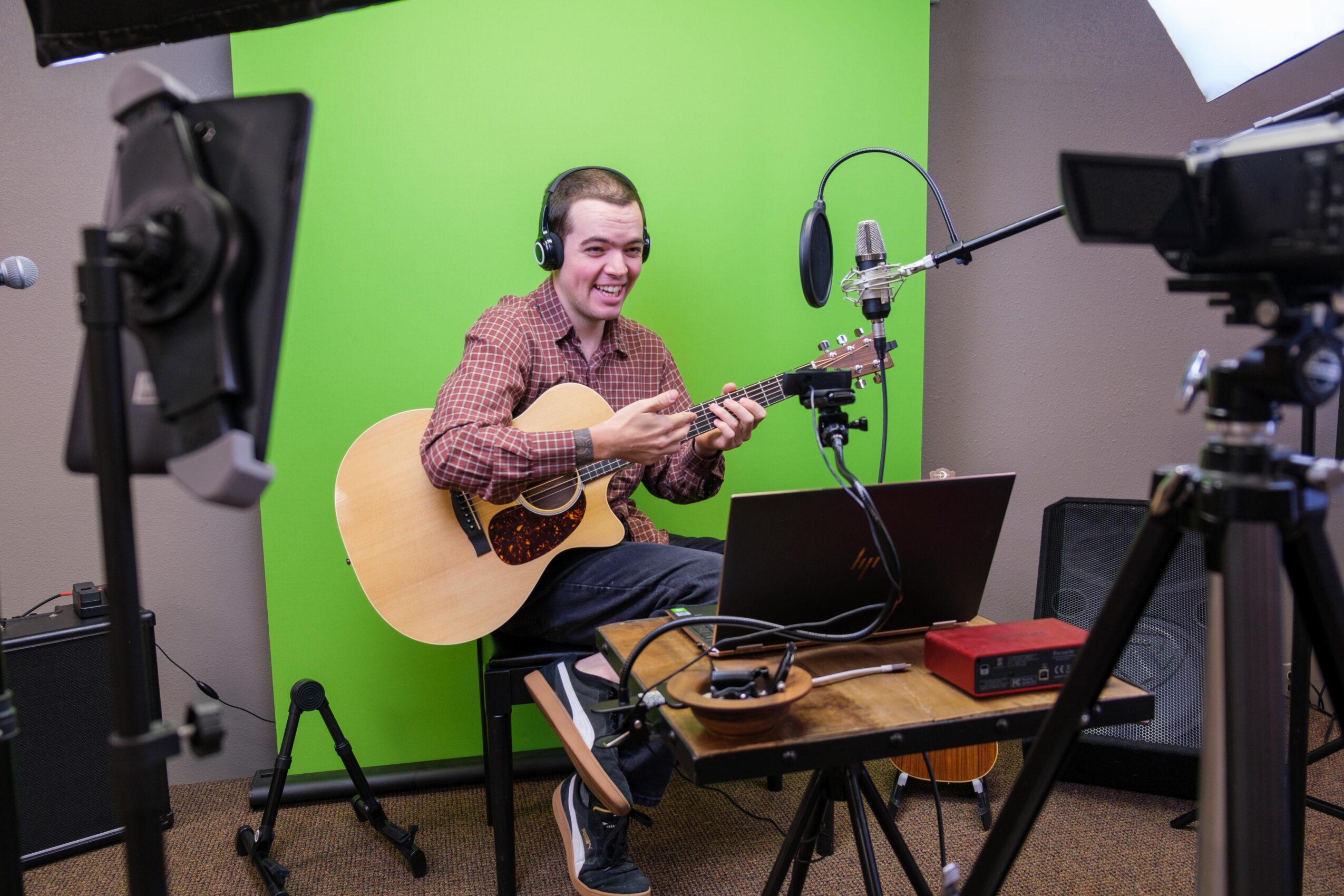 Person with headphones playing guitar in front of a green screen, surrounded by cameras and equipment, smiling at the camera.