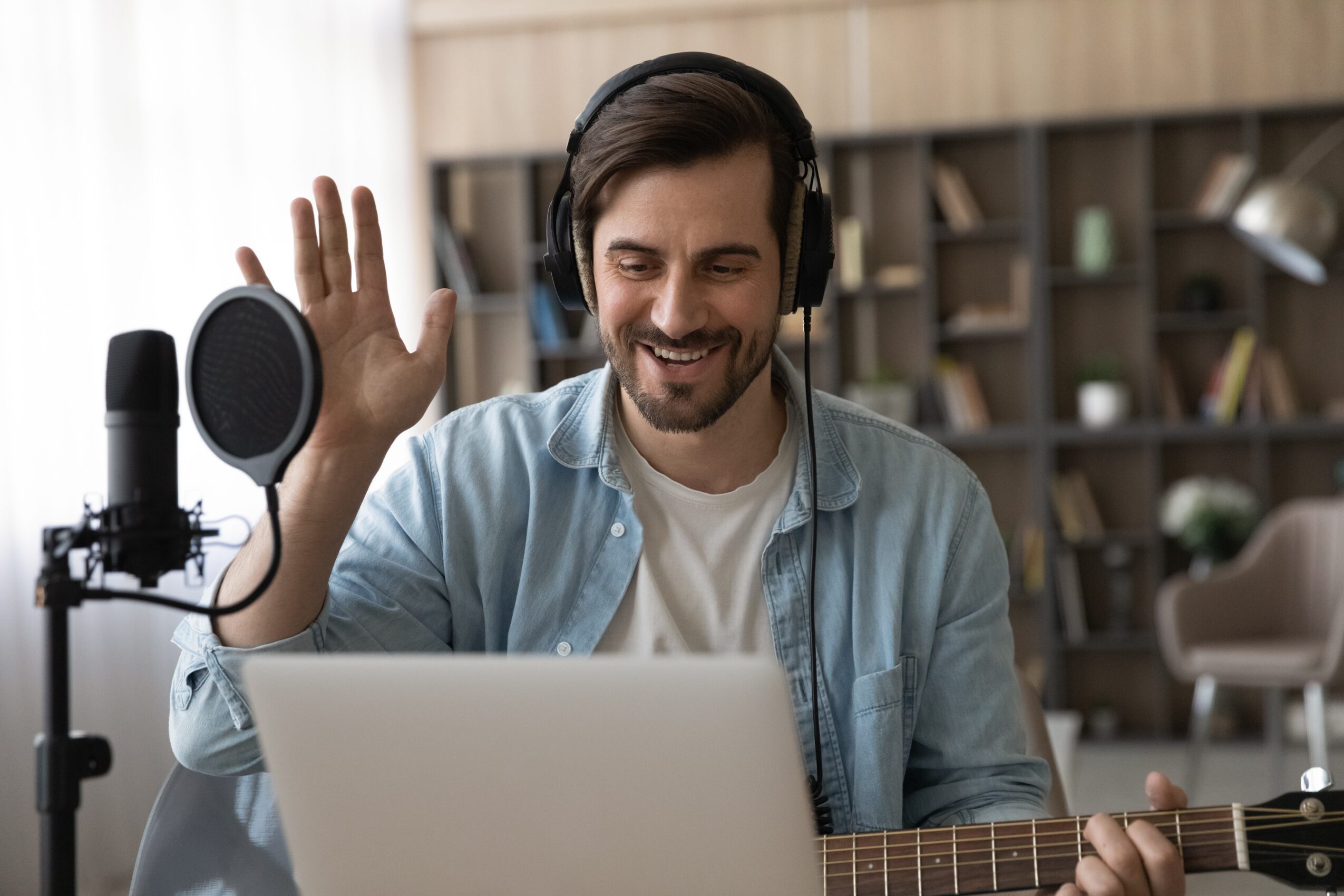 Man with headphones smiling and waving, sitting with a laptop and guitar in a room with bookshelves.