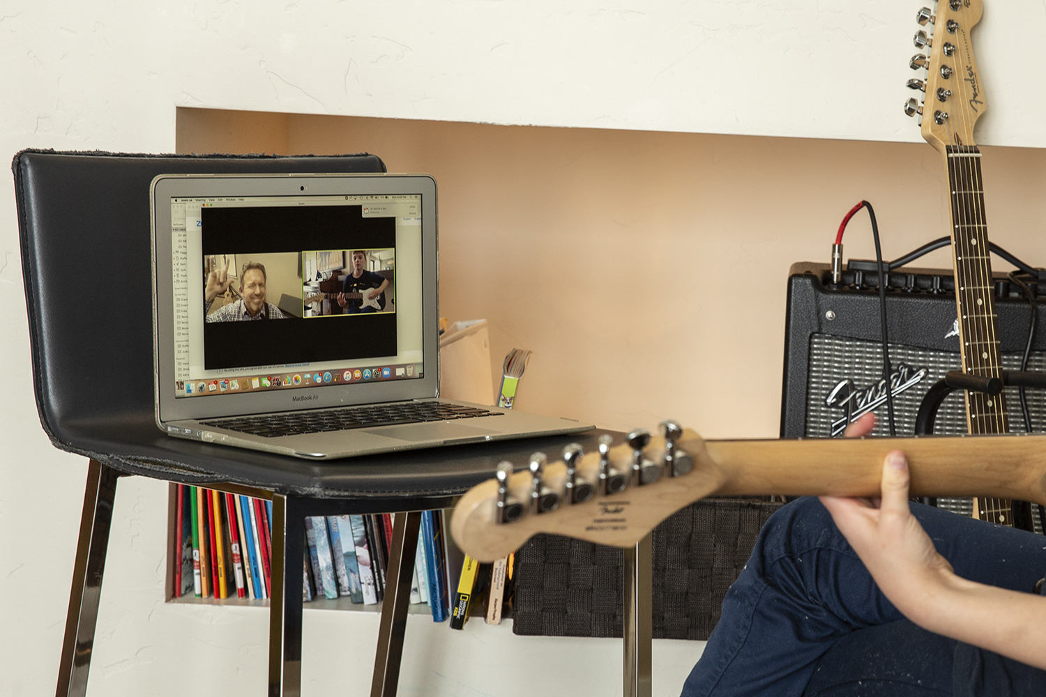 Person's hand holding a guitar neck, laptop on table showing a video call, amplifier, and books in background.
