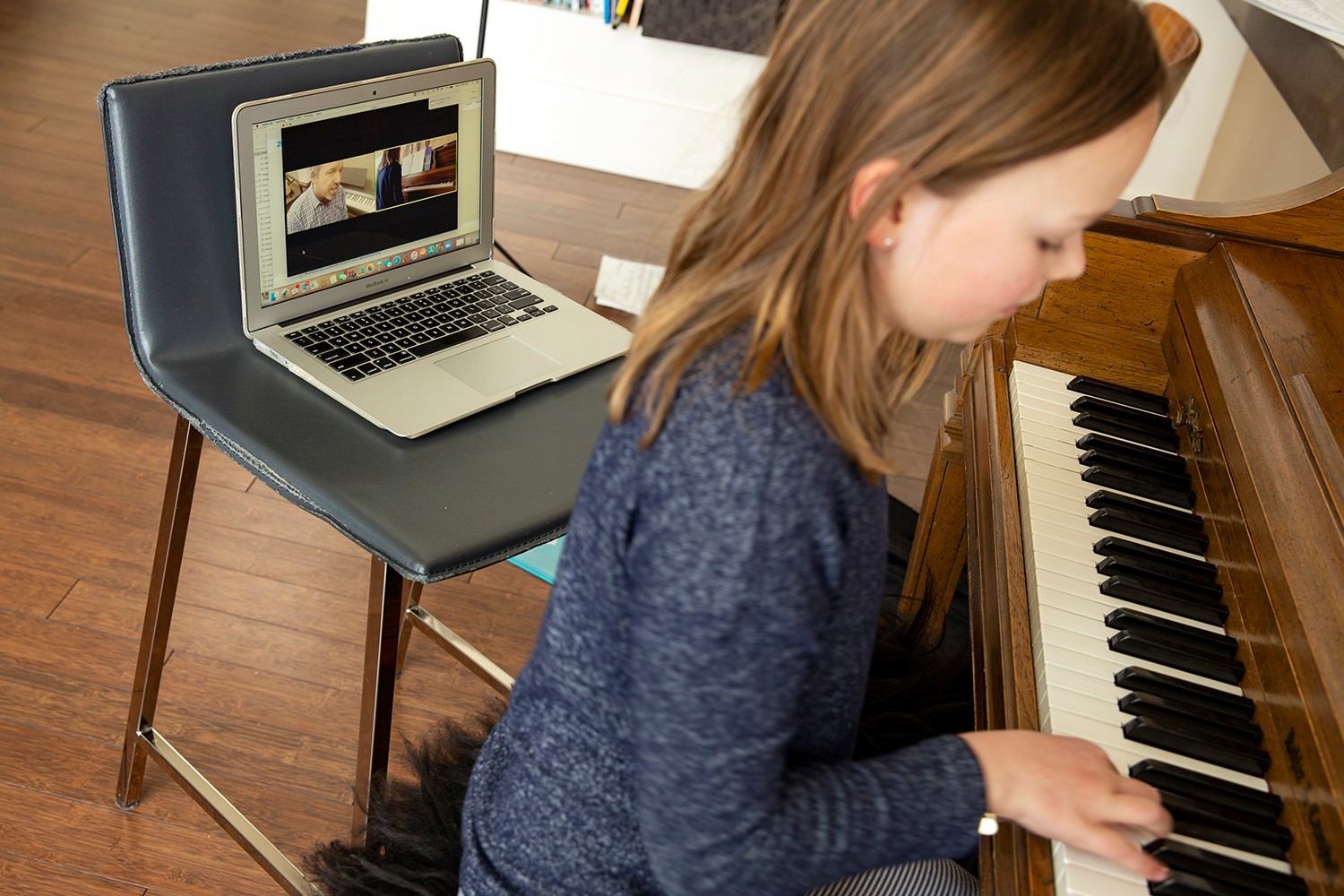 Woman playing piano with a laptop on a chair showing a video or image, in a room with wooden floor.