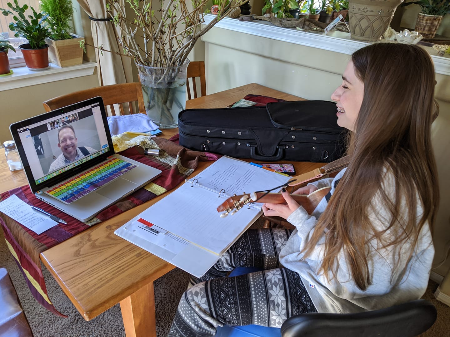 Young girl with long hair studying at a wooden table with a laptop, notebooks, and a backpack, in a bright room.