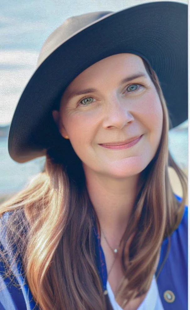 Woman with long hair wearing a wide-brimmed hat smiling outdoors, background of sky and water