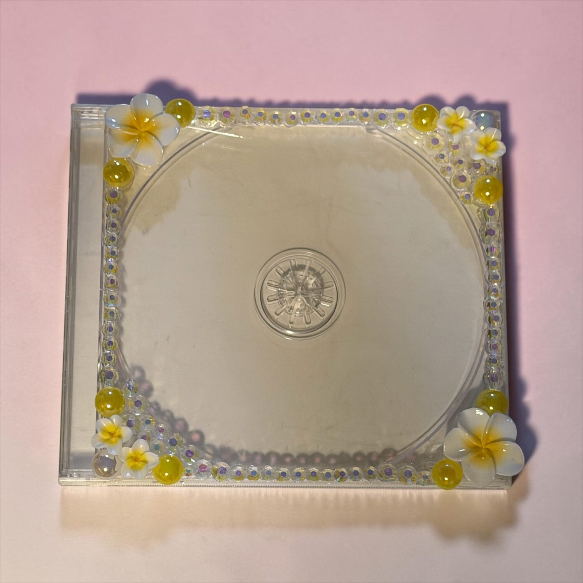 Clear square object with decorative flowers and beads on top, placed on a pink surface.