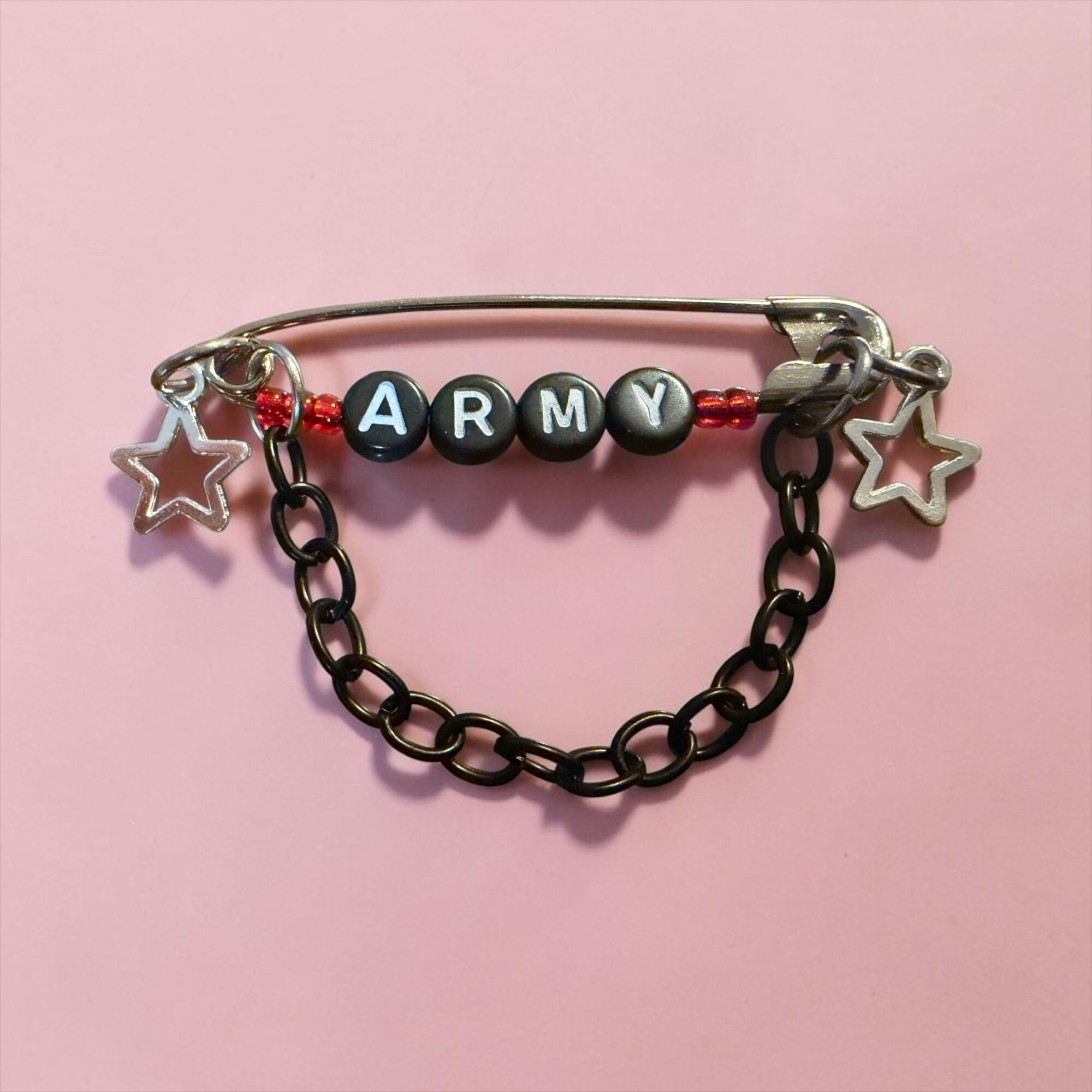 Decorative safety pin with beads spelling 'ARMY', star charms, and a chain, on a pink background.