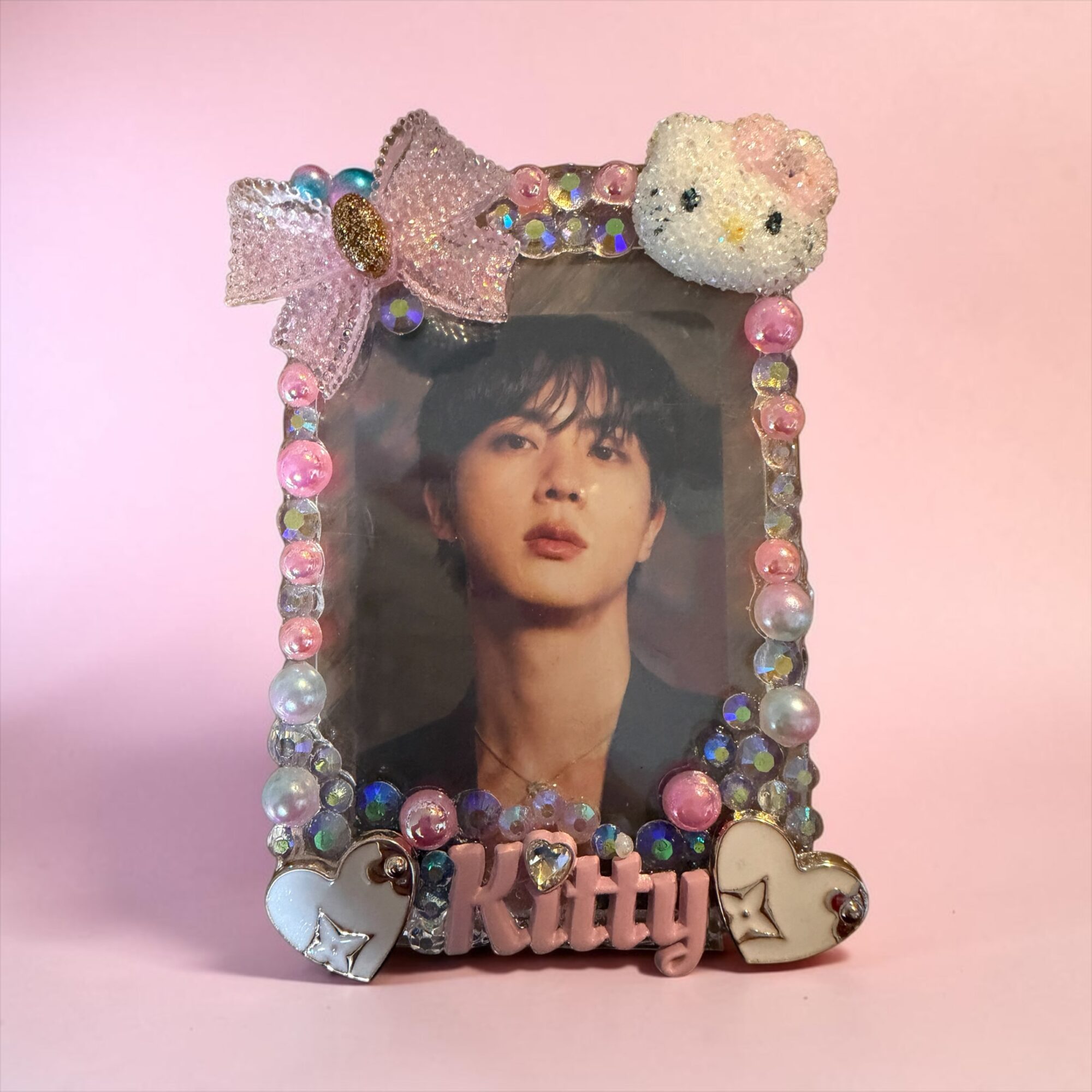 Decorative photo frame with a woman's portrait, adorned with a bow, a bear, and beads, against a pink background.