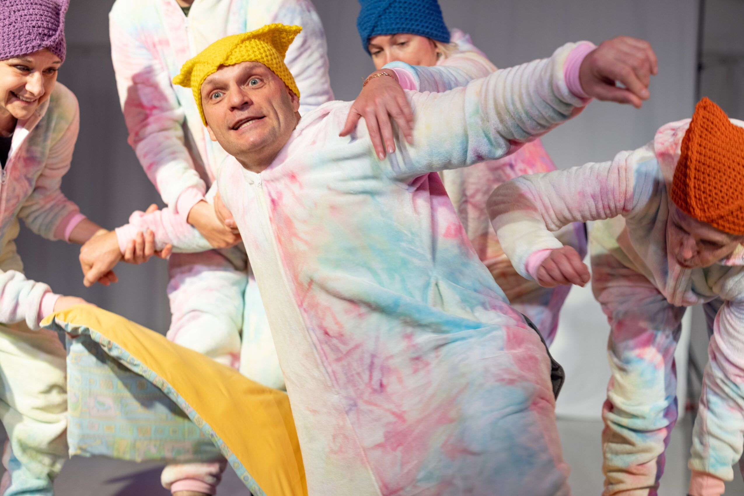 Group of people wearing colorful, tie-dye outfits and knit hats, holding hands and dancing together indoors.