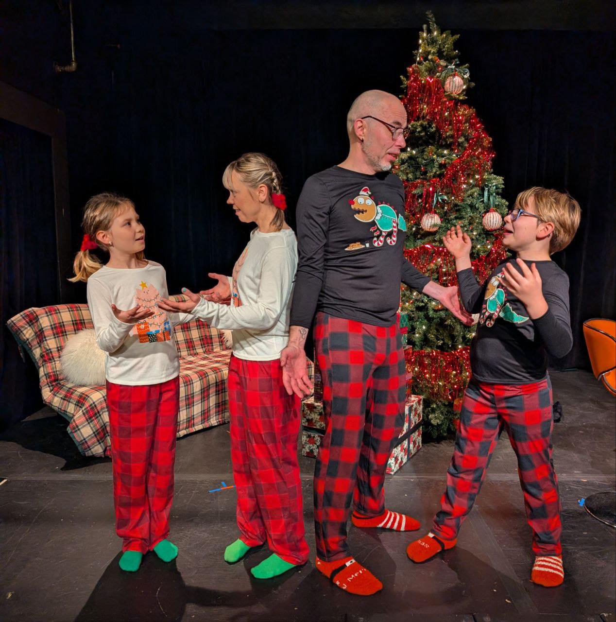 Four people in Christmas pajamas stand near a decorated tree, engaging in conversation, with a couch and holiday decorations in the background.