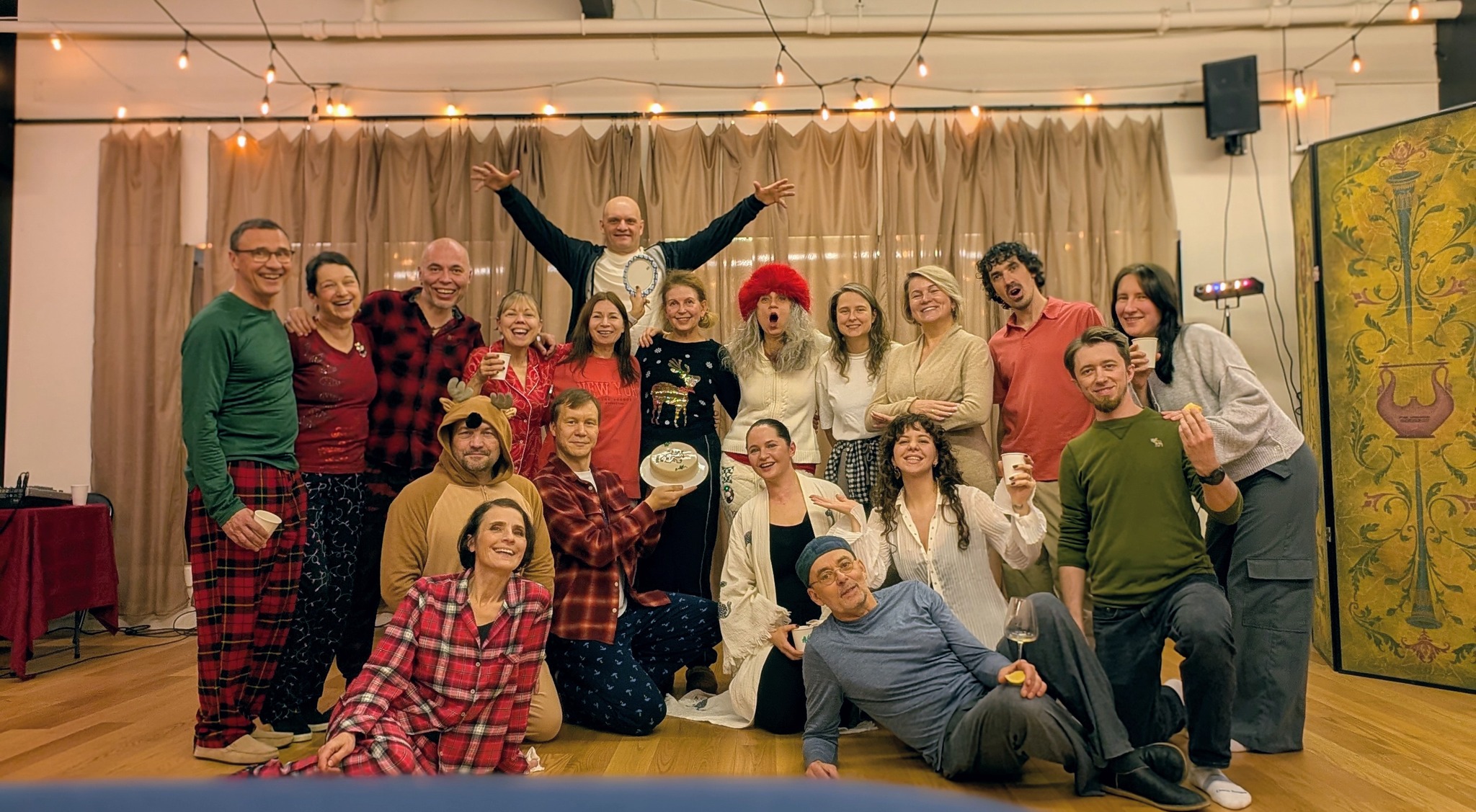 Group of people posing indoors with a curtain backdrop, some holding drinks, smiling, and dressed casually.