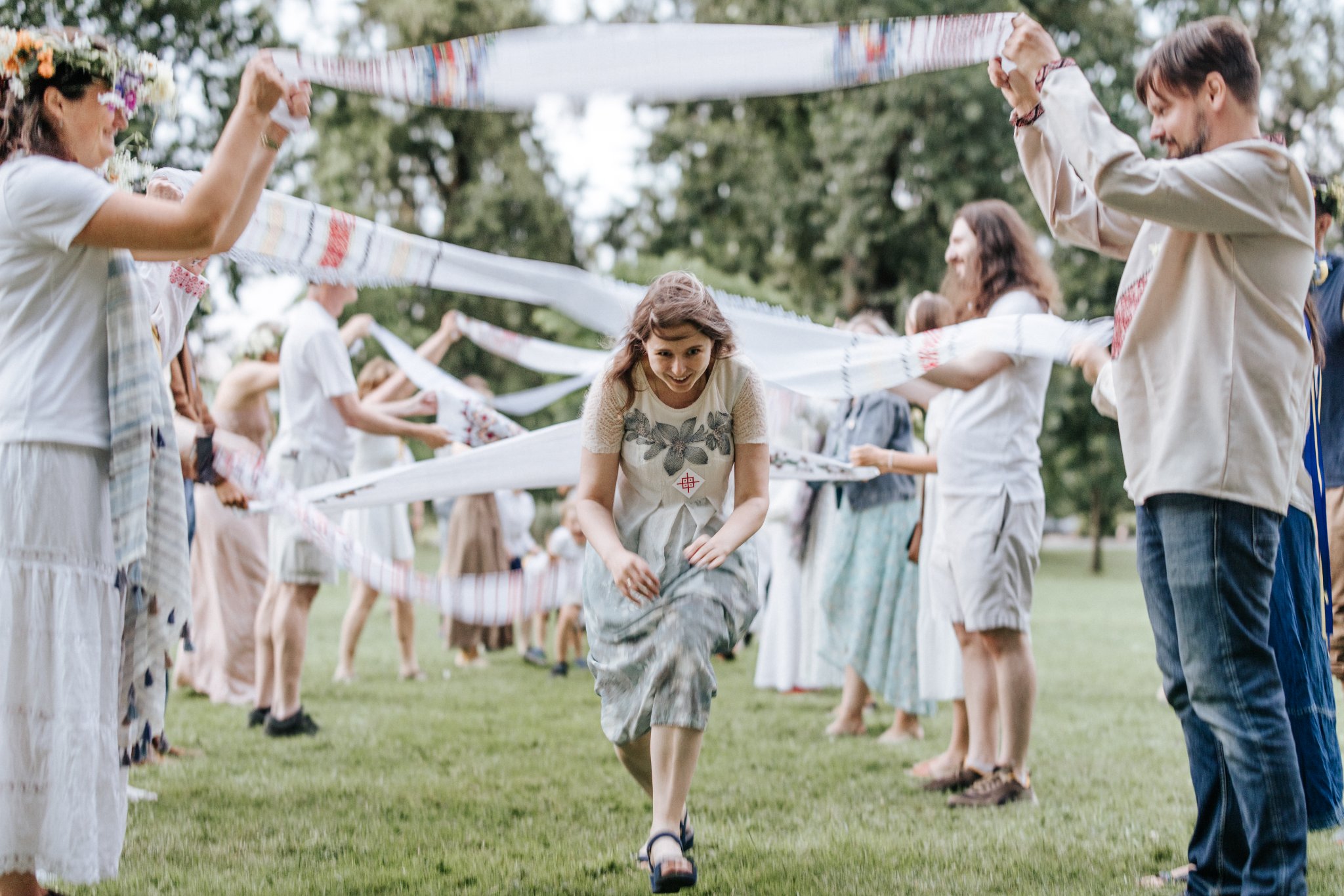 Group of people outdoors holding fabric strips, forming a circle around a girl in a dress, on a grassy field.
