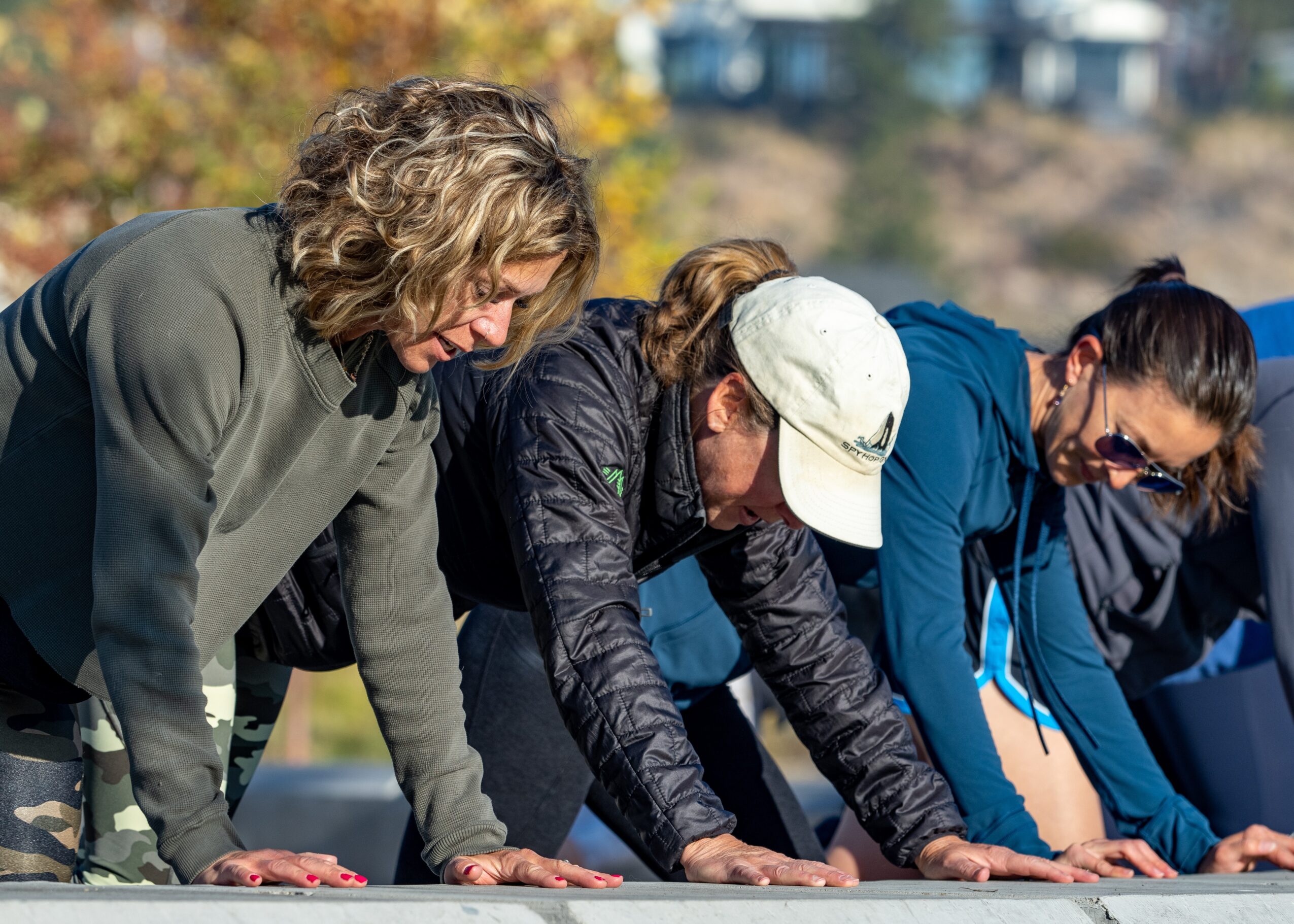 Three women are doing push-ups outdoors on a sunny day, with trees and buildings in the background.
