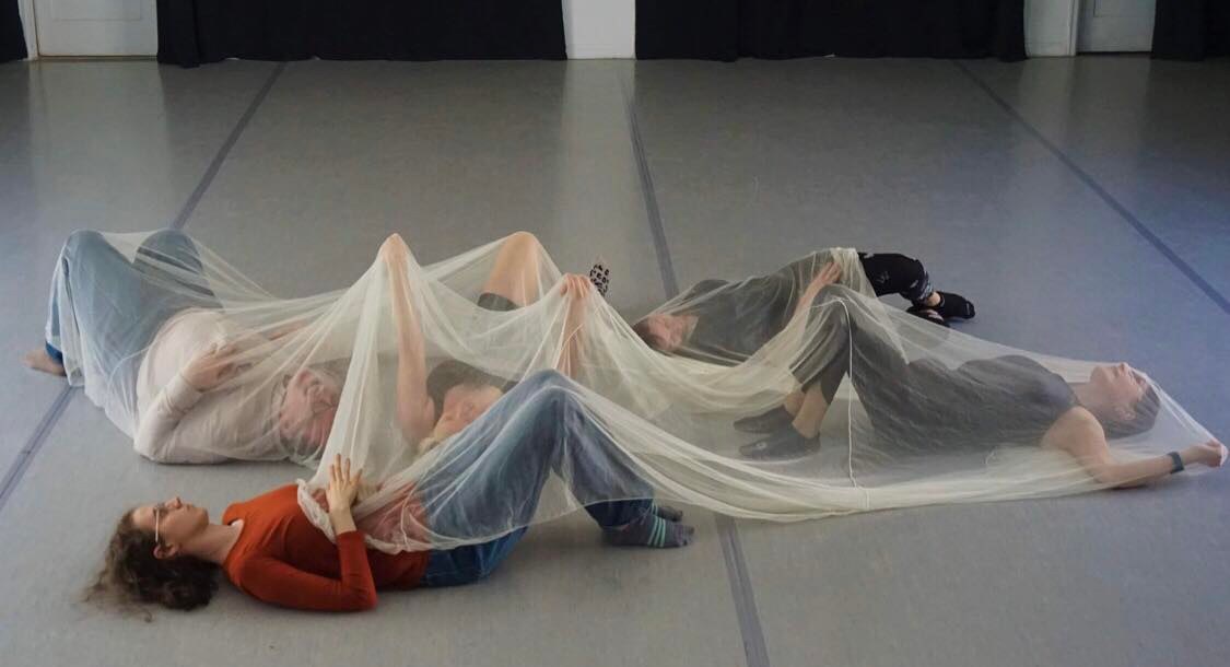Four people lying on the floor under a sheer fabric, with their heads and feet visible, in a dance or performance setting.